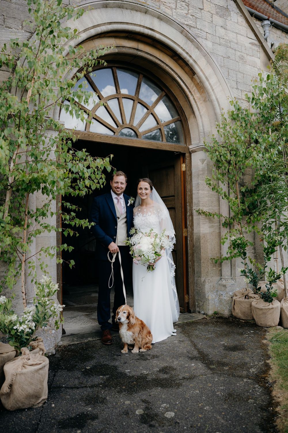 Bride and groom stand with their Basset Hound dog in a stone archway doorway decorated with greenery.