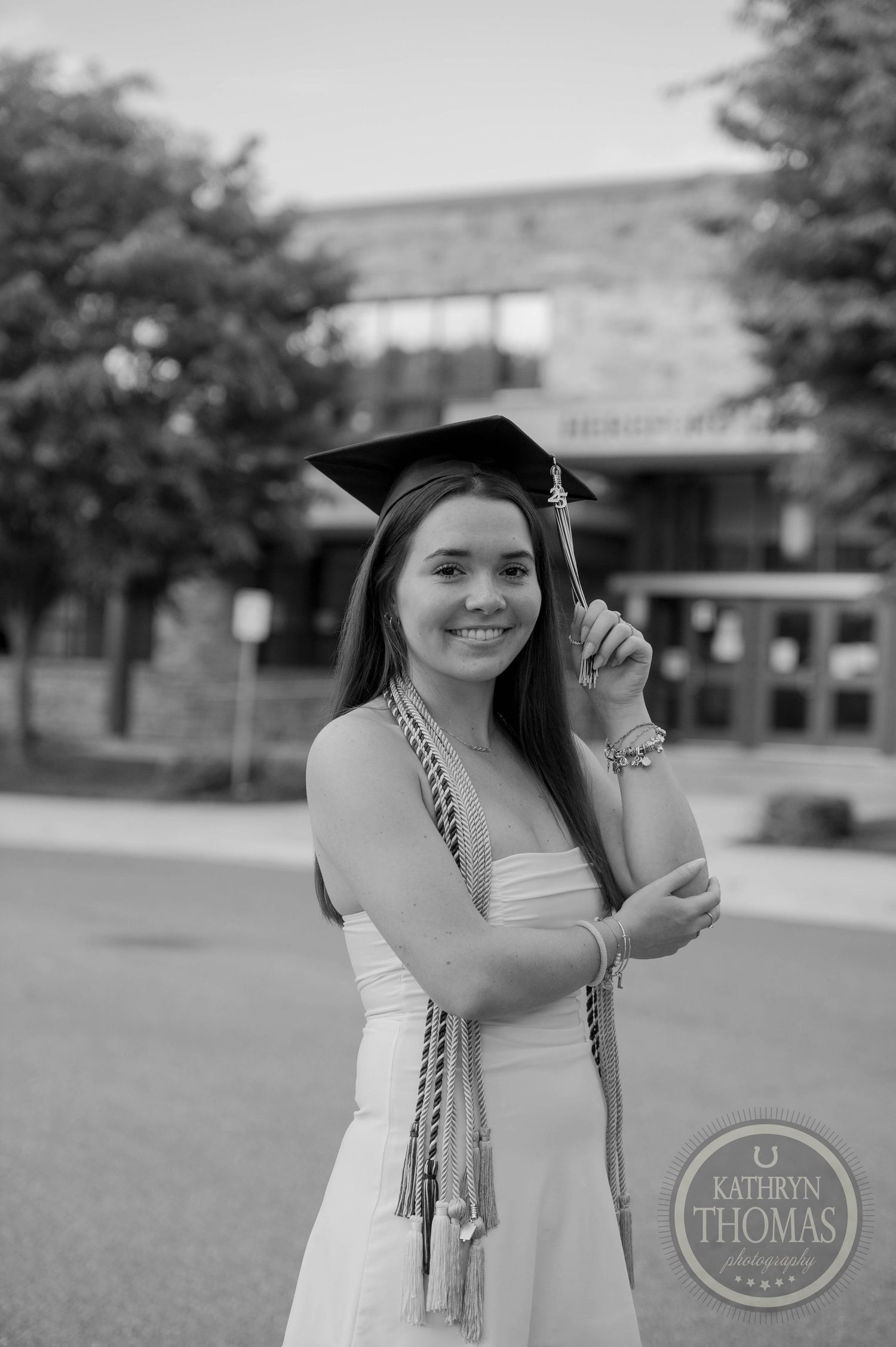 Graduate in white dress and gold honor cords poses outdoors with graduation cap and academic regalia.