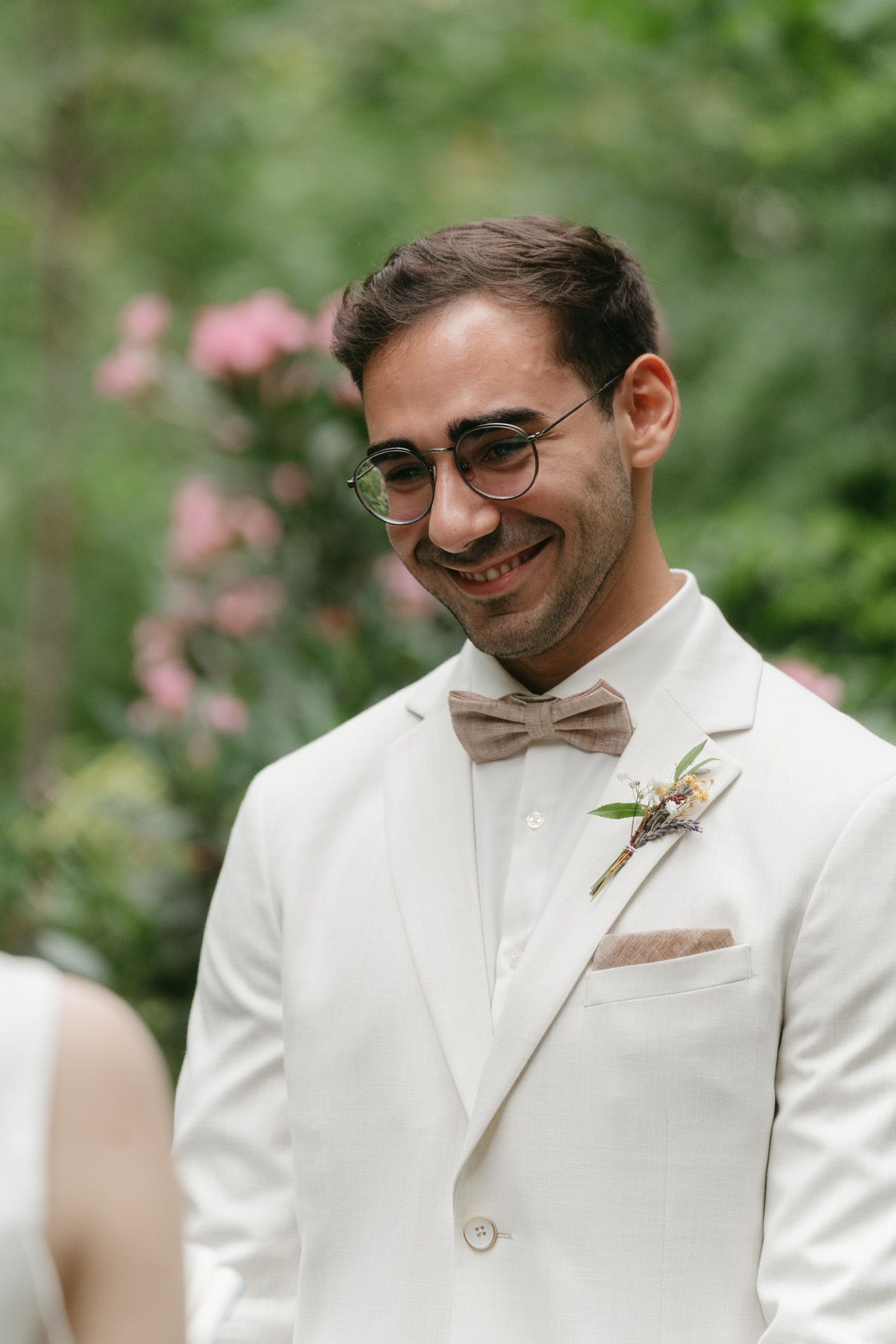 A groom in a white suit and bow tie smiles in a garden setting.