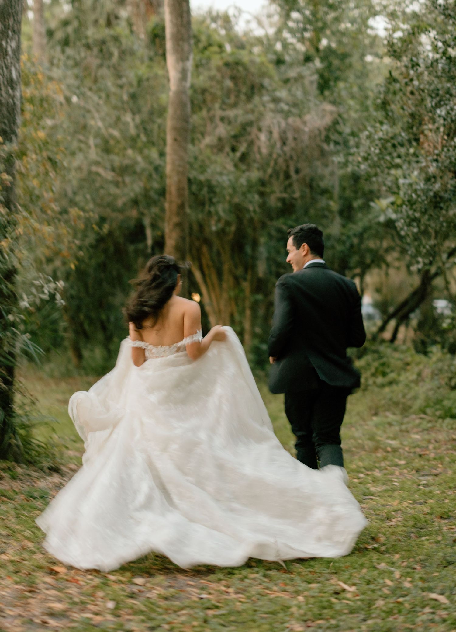A cinematic photo of a couple in wedding attire runs playfully through a wooded area with flowing white dress trailing behind them.