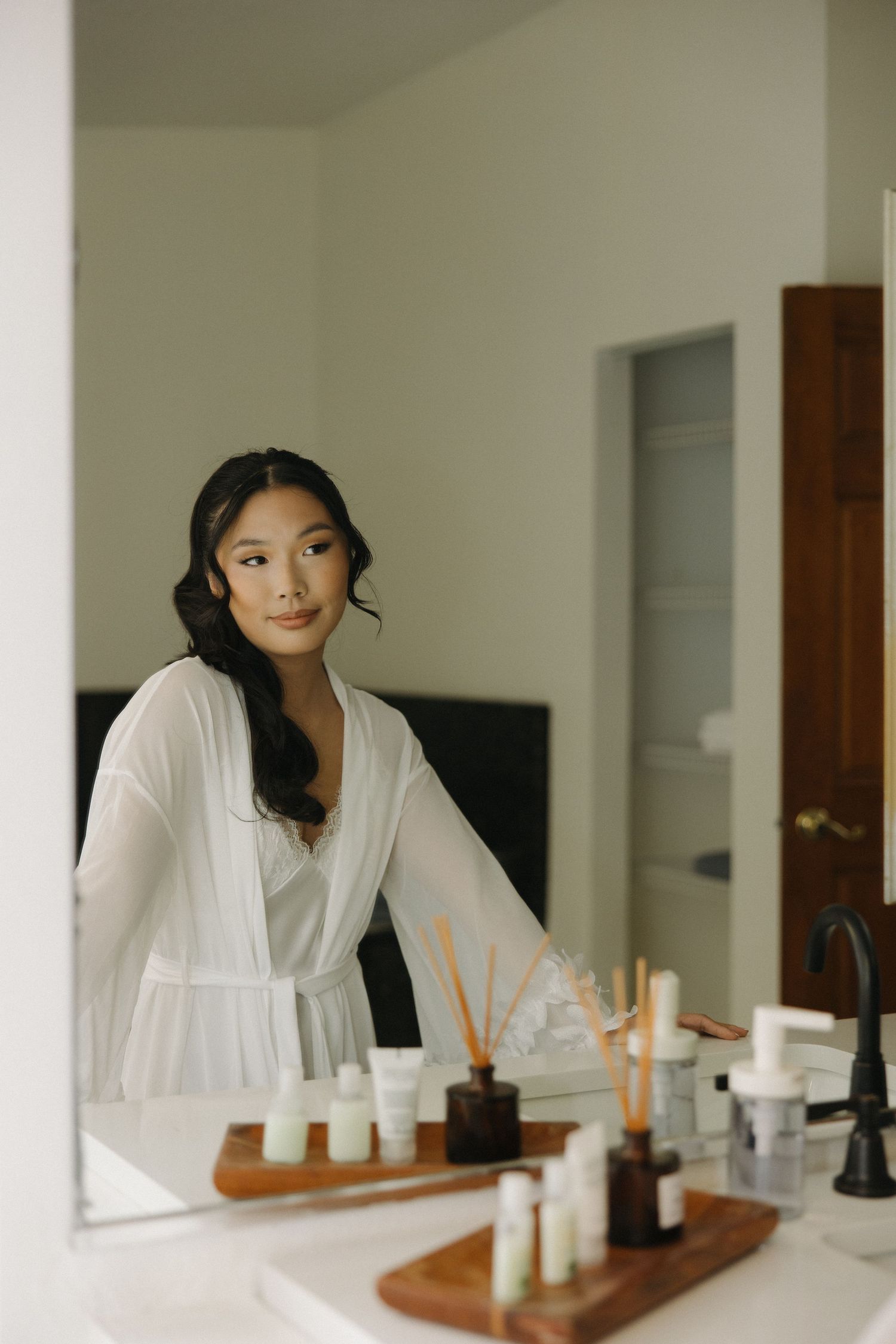 Person in white robe sits at vanity table with beauty products and candles in minimalist interior setting.