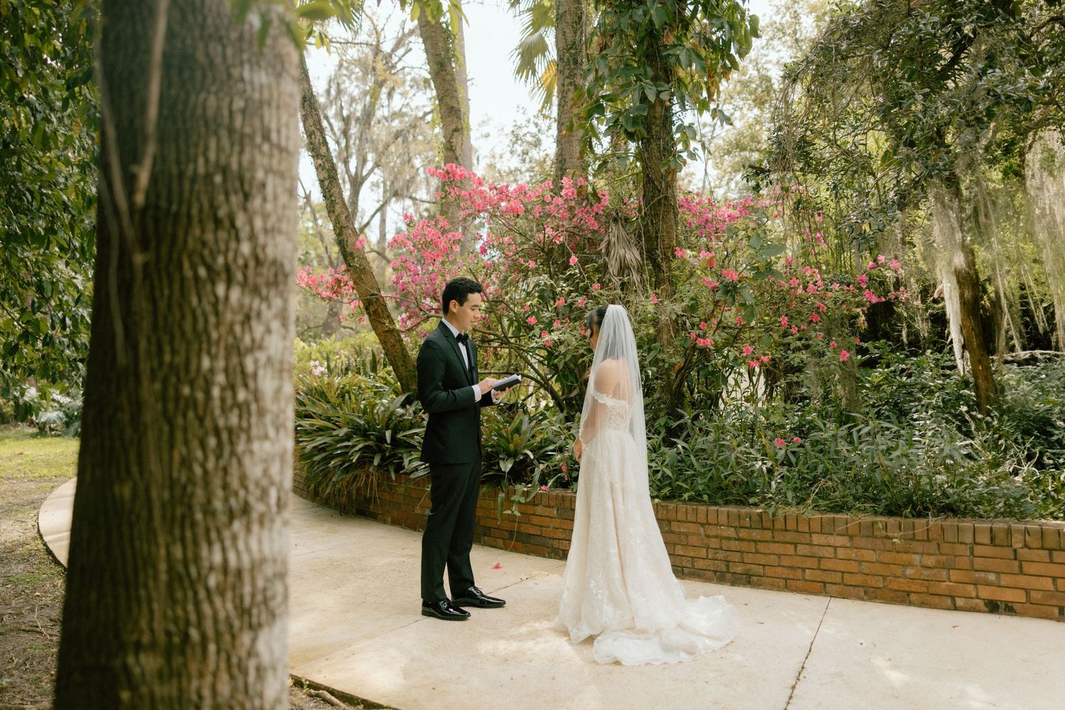 A couple in wedding attire shares a tender first look moment in a lush garden setting with tall trees.