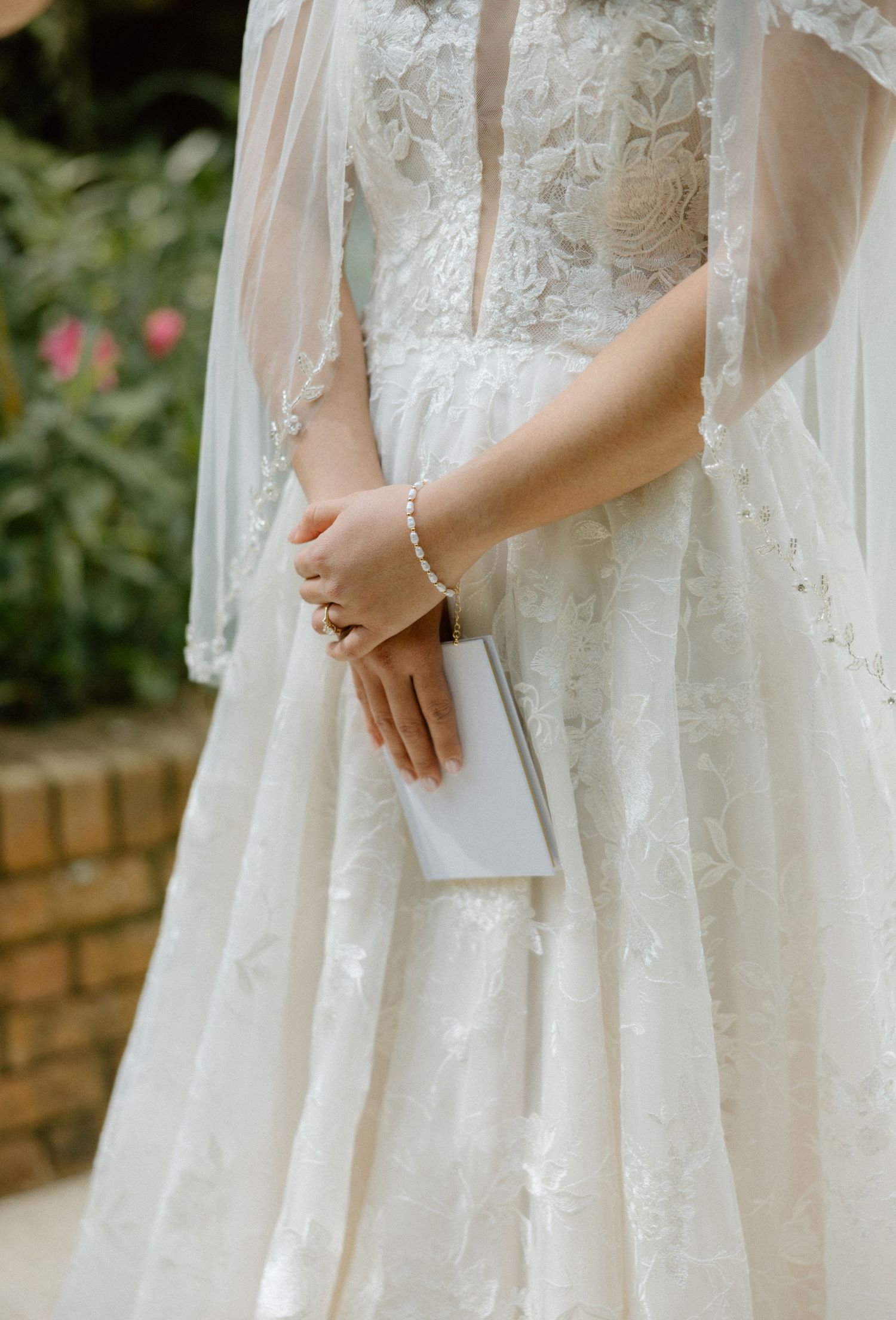 Close-up of a bride's hand holding a white clutch purse, wearing a delicate bracelet and lace wedding gown.