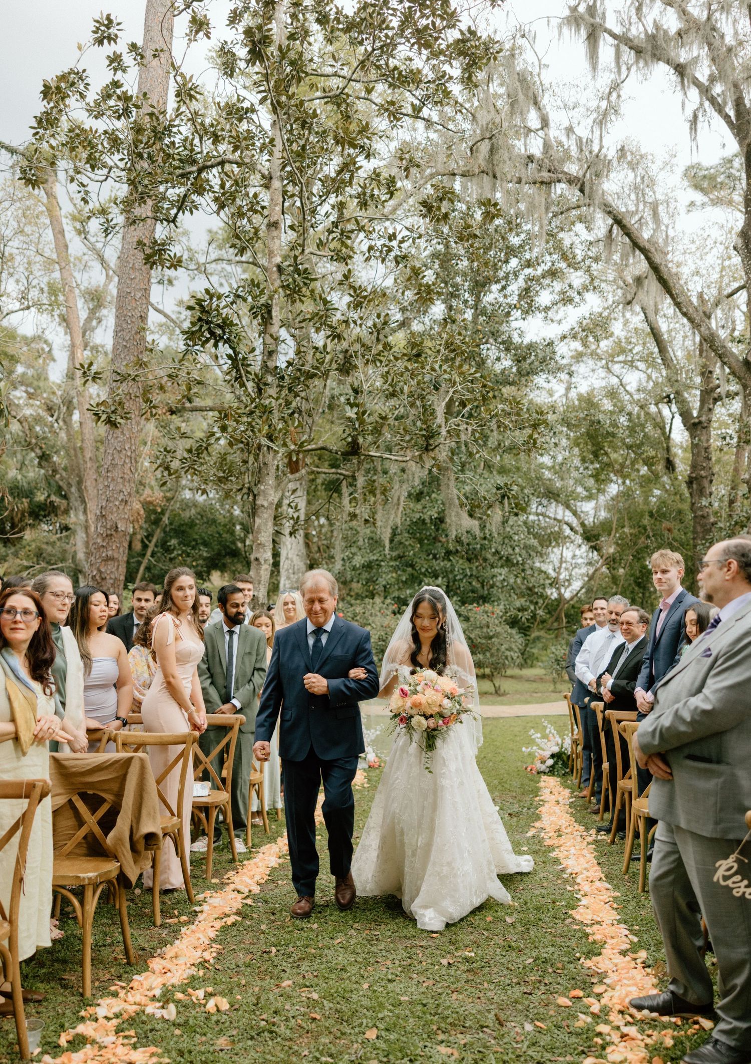 Outdoor wedding ceremony with wooden chairs and rose petal aisle lined by trees draped in Spanish moss.