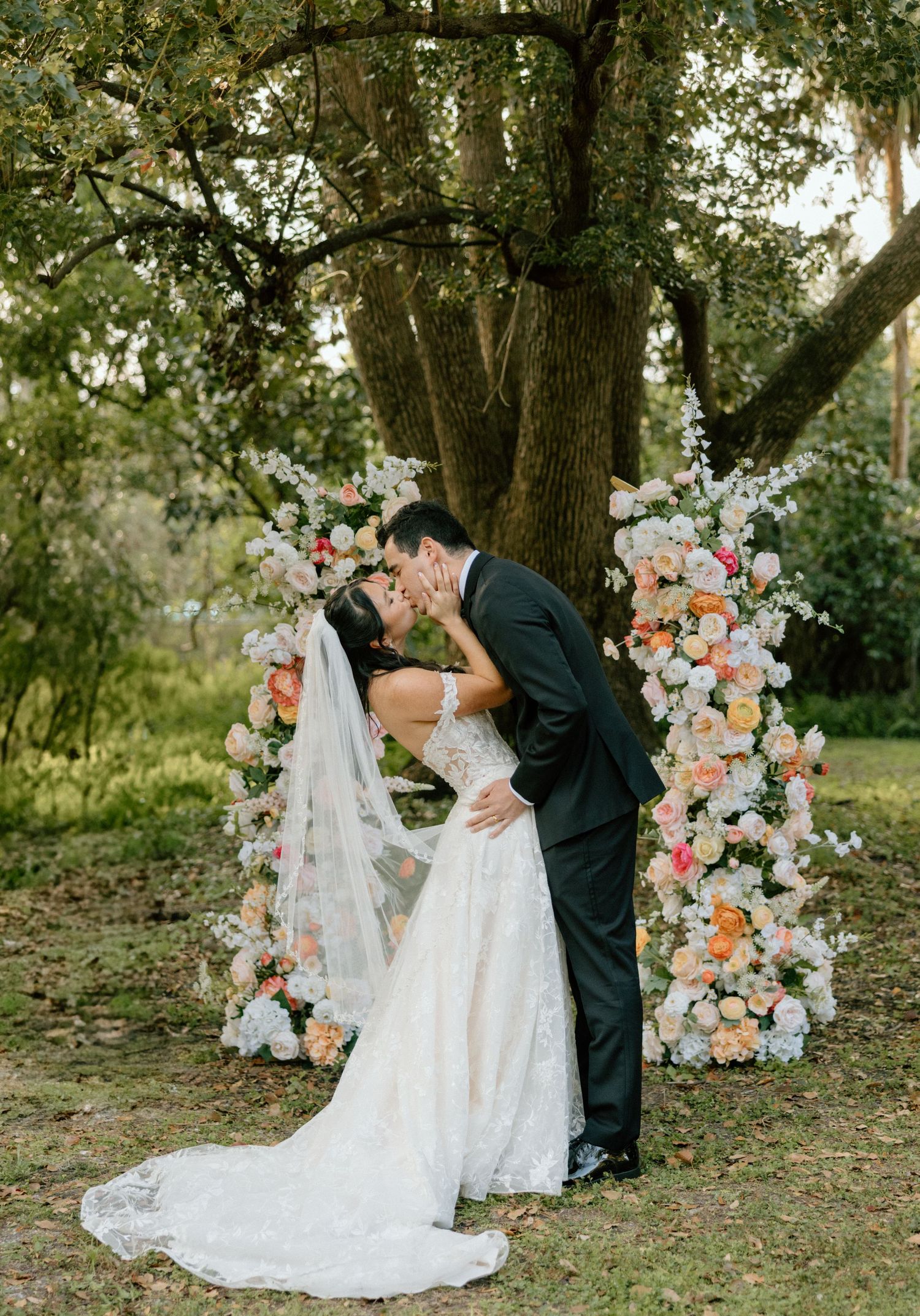Romantic wedding couple sharing a kiss by a floral arch with peach and white blooms in a garden setting.