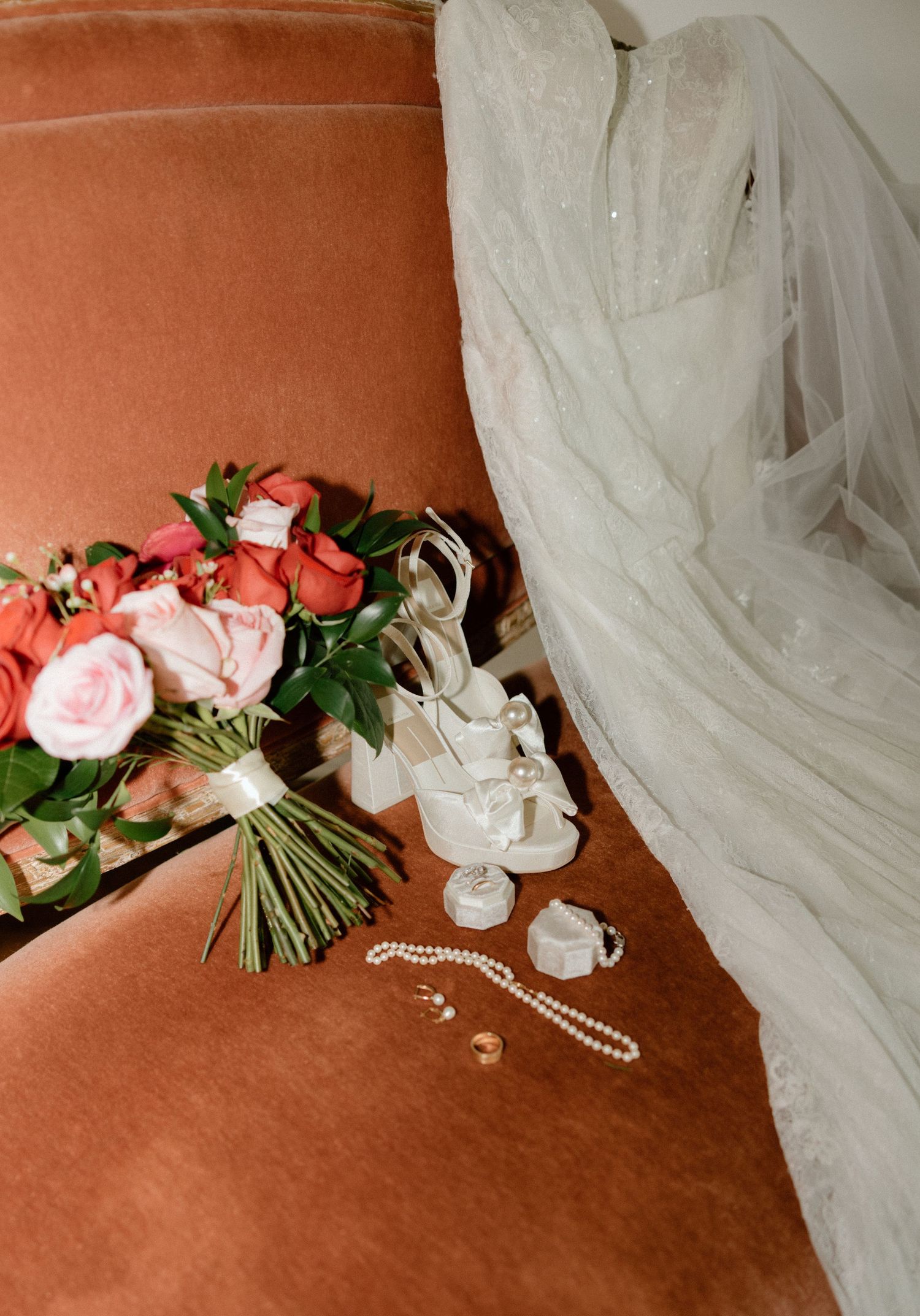 Wedding accessories including pink roses, white veil, and pearl necklace arranged on a terracotta colored surface.