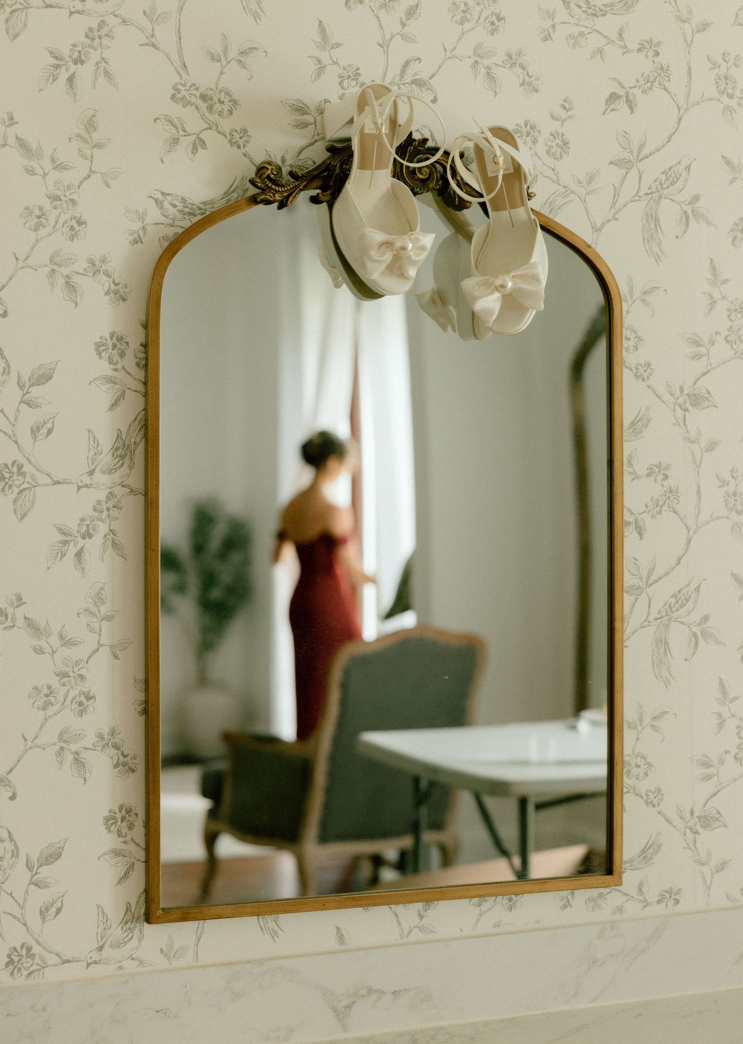 A vintage gold-framed mirror hangs on floral wallpaper, with a blurred reflection showing a red silhouette in the background.