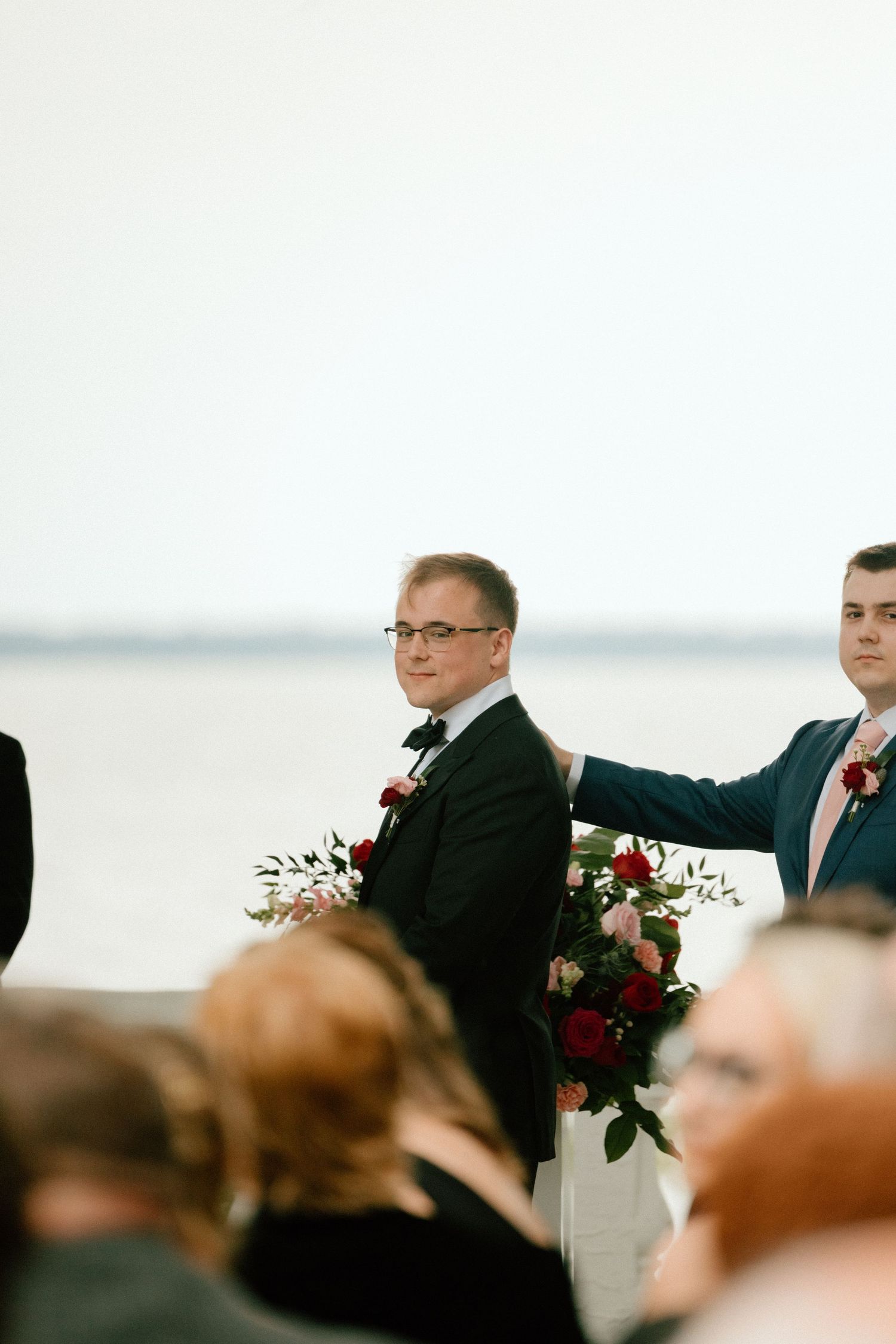 The groom during the wedding ceremony by the water with guests watching and red and white floral arrangements decorating the scene.