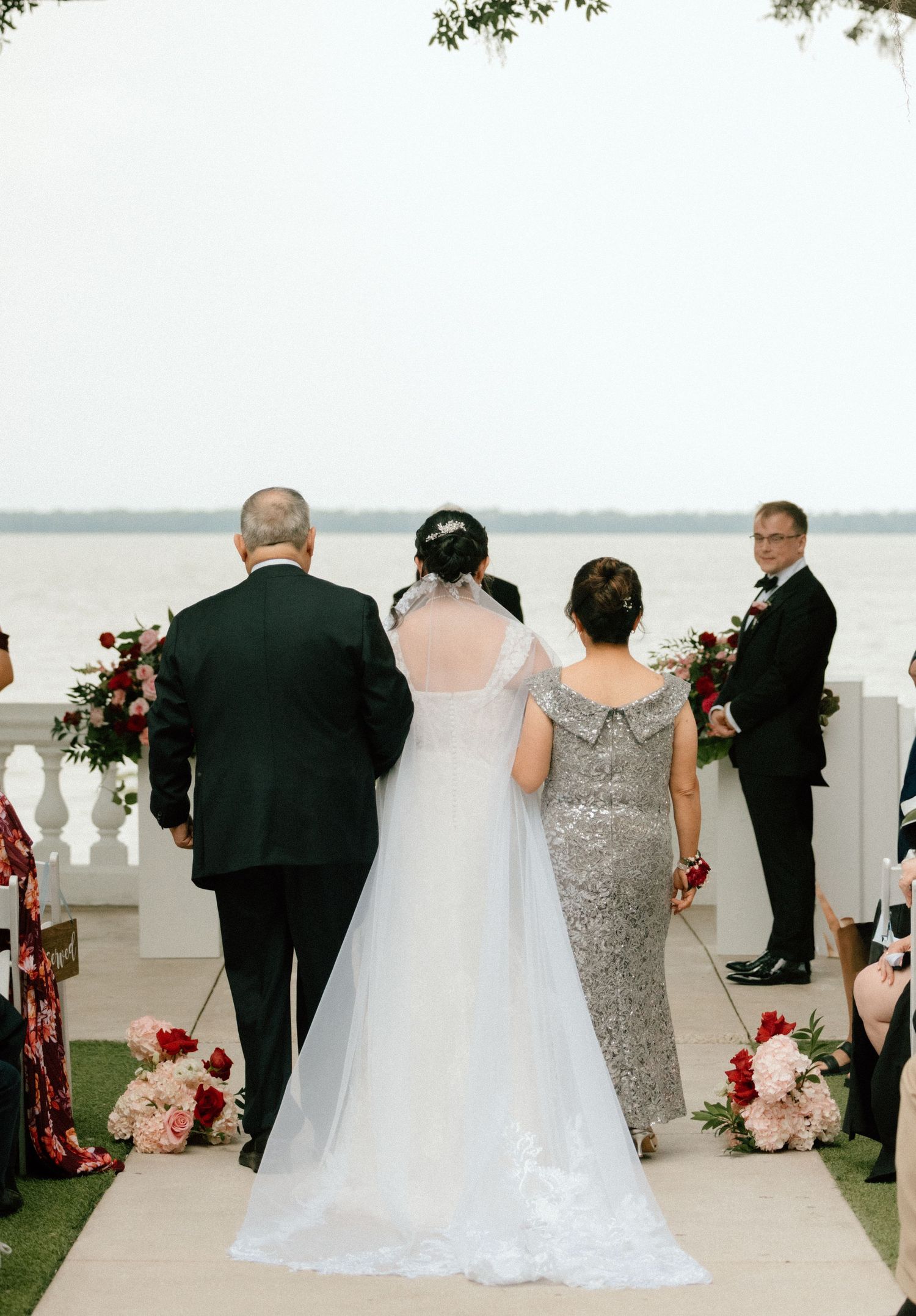 Wedding party gathered on a waterfront terrace with pink and red floral decorations.