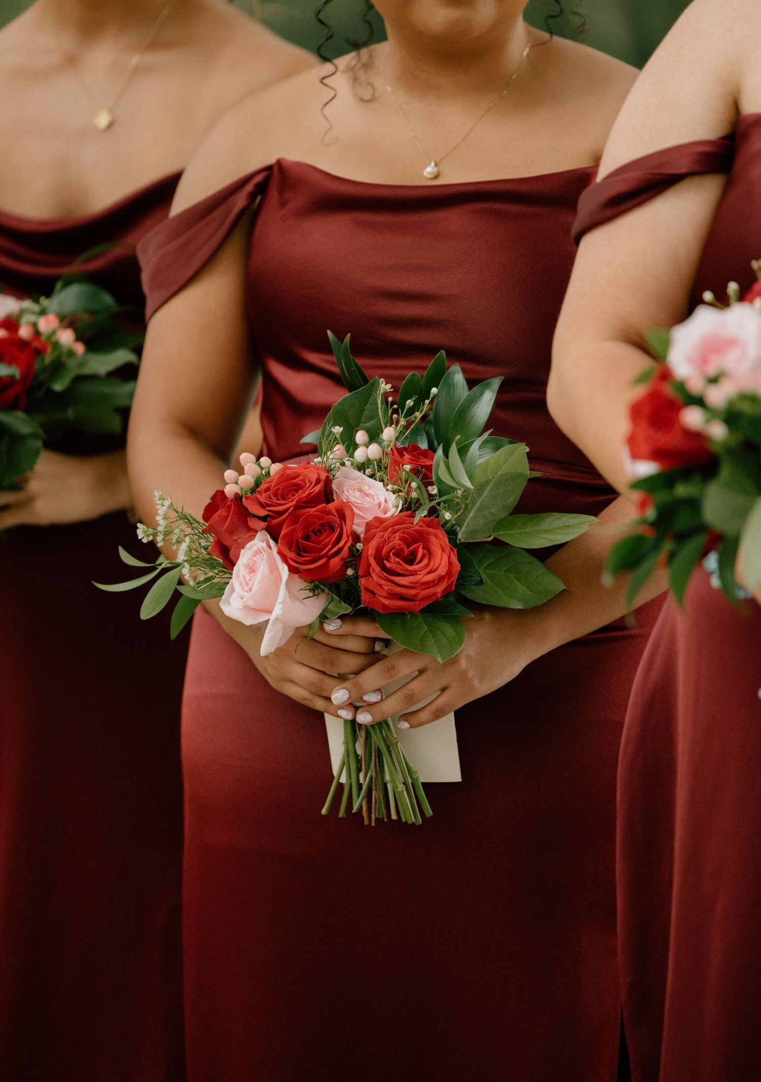 Bridesmaid in burgundy off-shoulder dress holds elegant bouquet of red and pink roses with greenery.