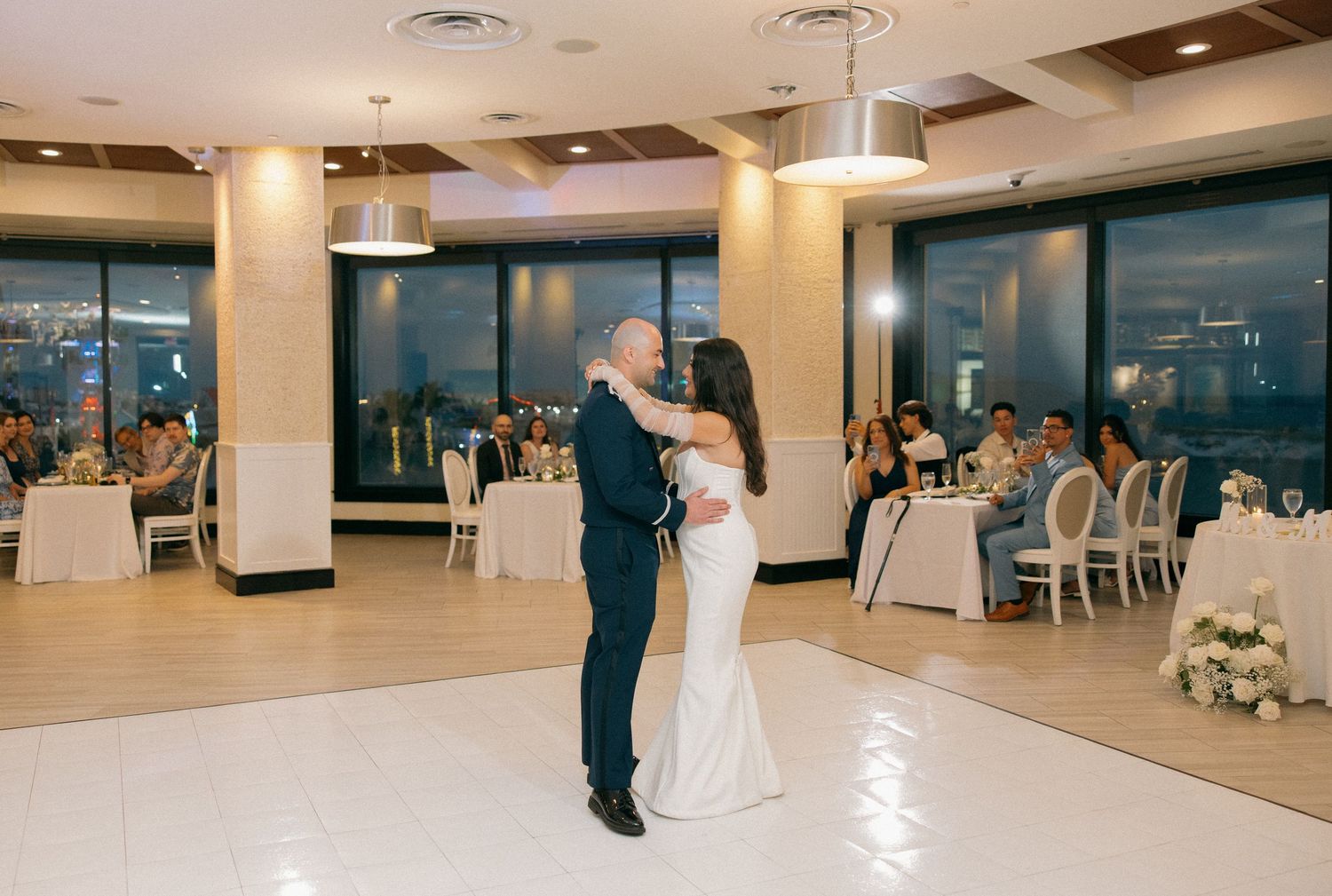 Couple sharing their first dance together in an elegant white reception hall with modern lighting and decor.