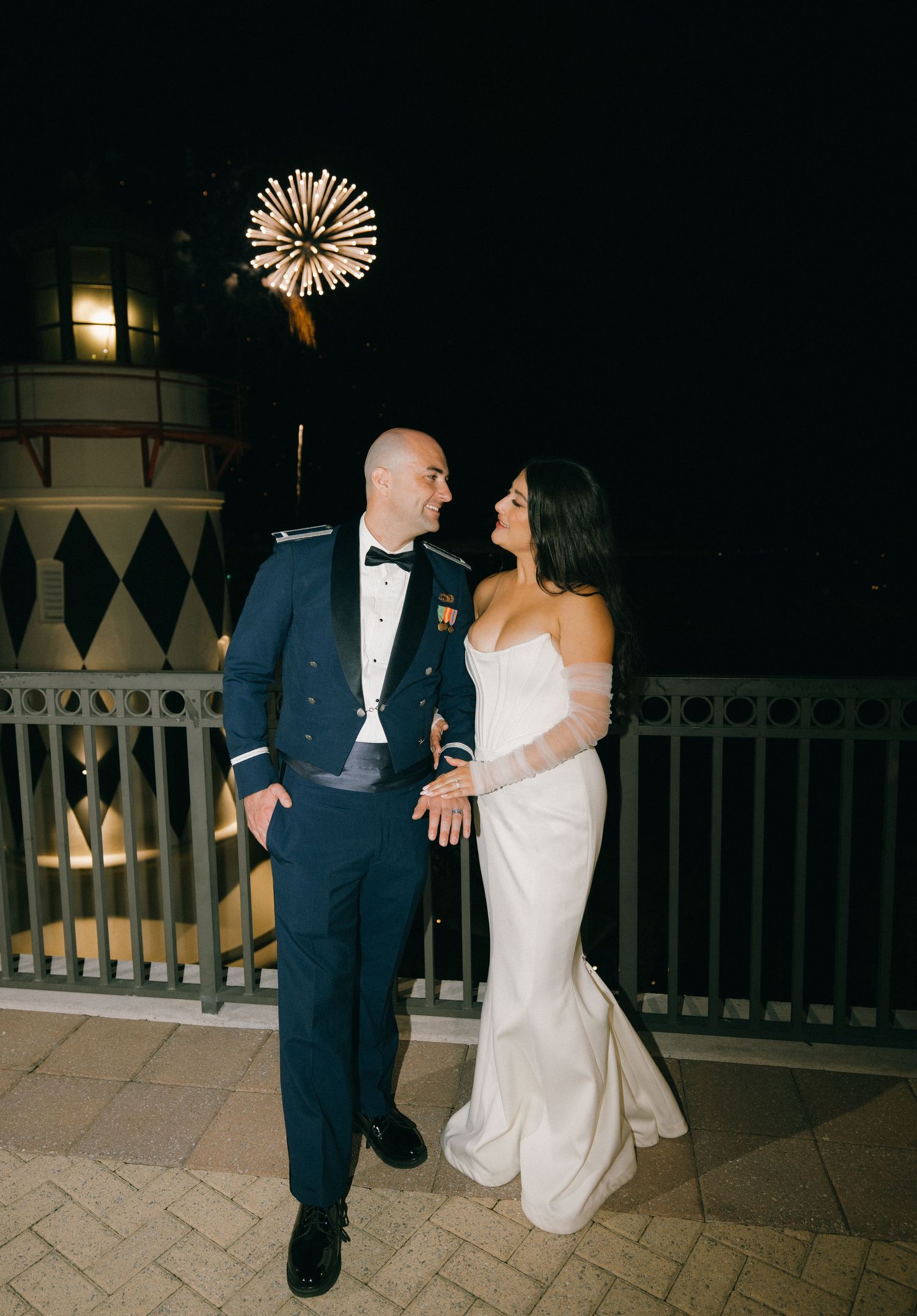 A couple in wedding attire shares a romantic dance moment on a lit patio at night with fireworks in the background.