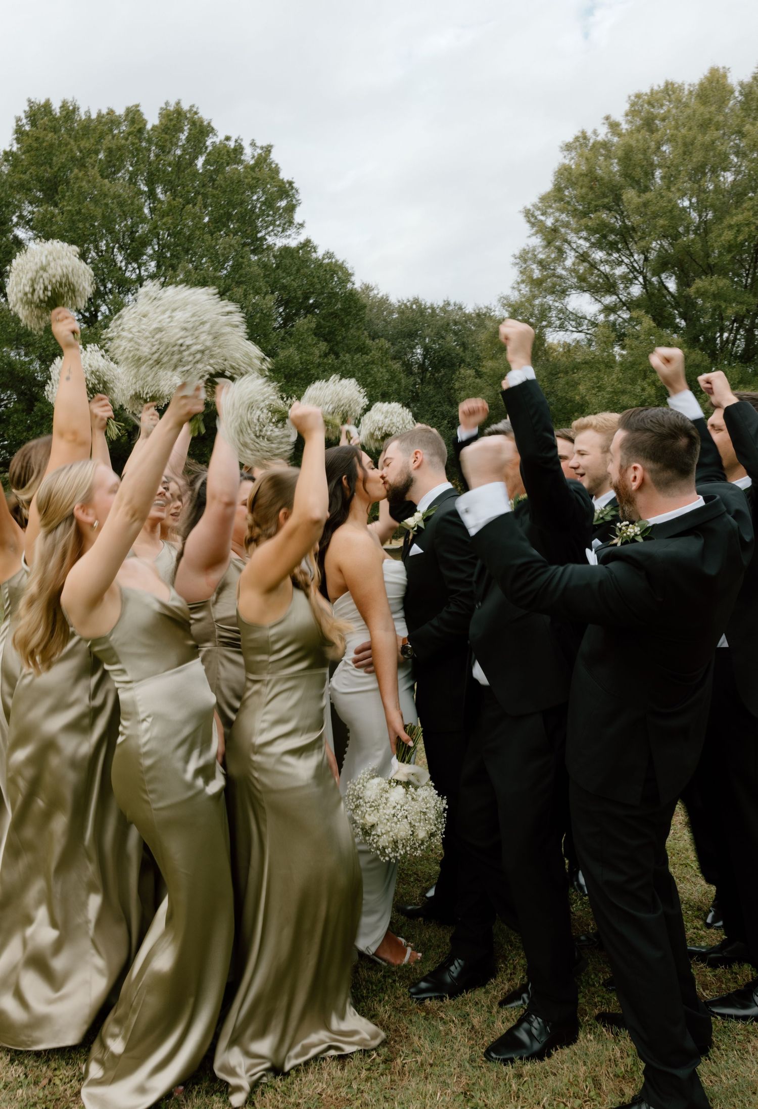 Wedding party members in black suits and champagne dresses turn their backs to camera and raise their arms in celebration.