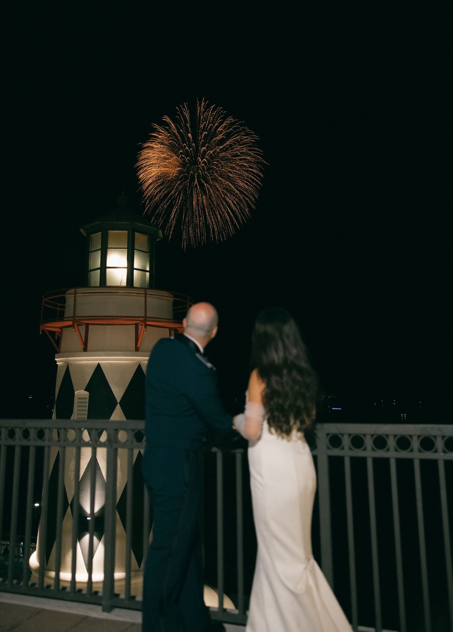 A couple watches fireworks burst over a lighthouse at night from a railing deck during a romantic evening.