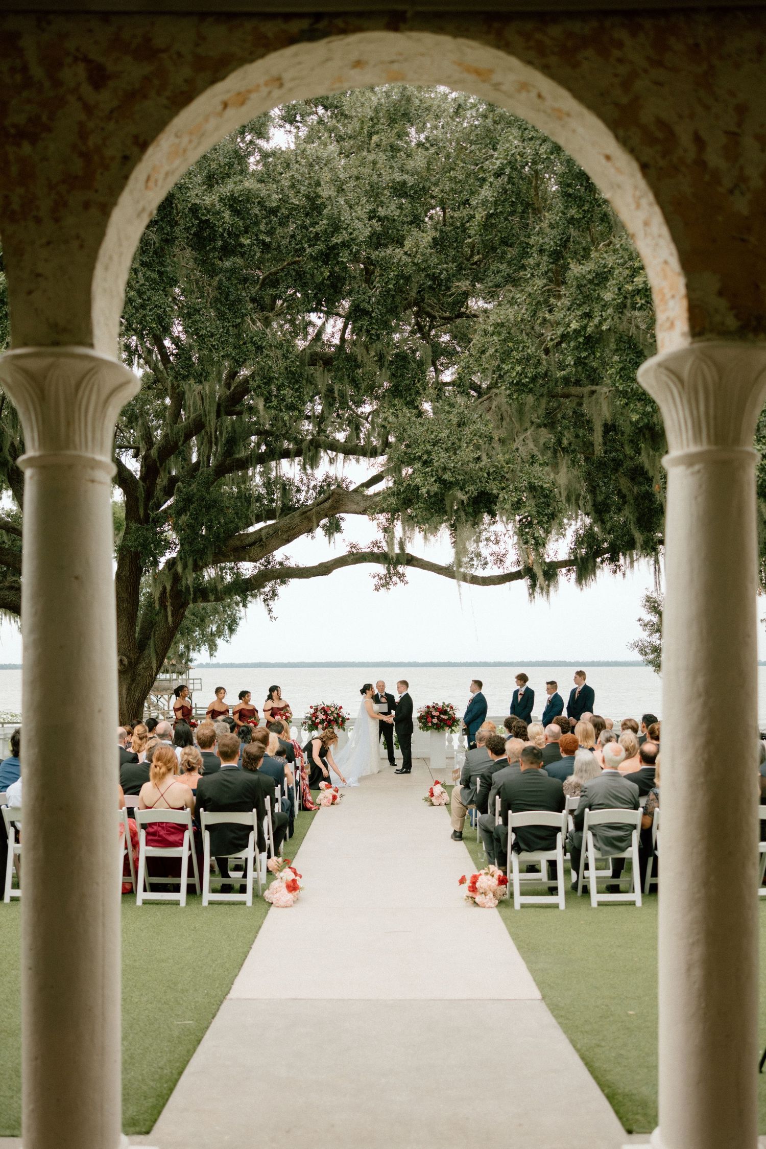 Wedding ceremony setup with white chairs under arched columns overlooking a waterfront view.
