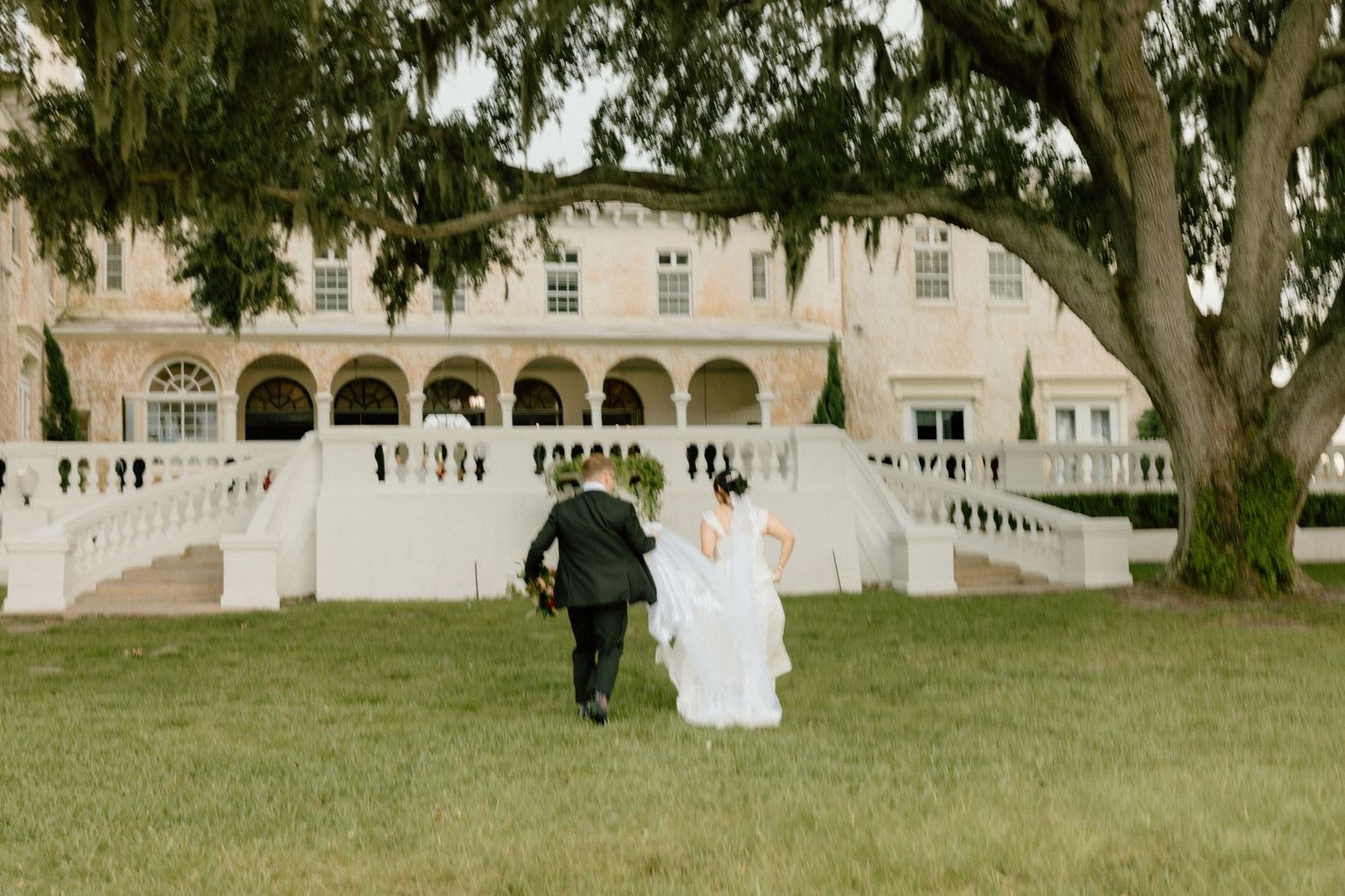 Wedding guests gather on a grand estate lawn with elegant white architecture and mature trees in the background.