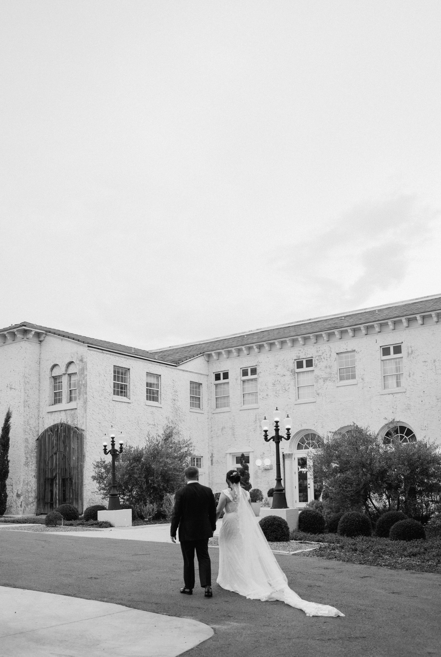 A couple in wedding attire walks along a path outside an elegant Mediterranean-style building with ornate architecture.