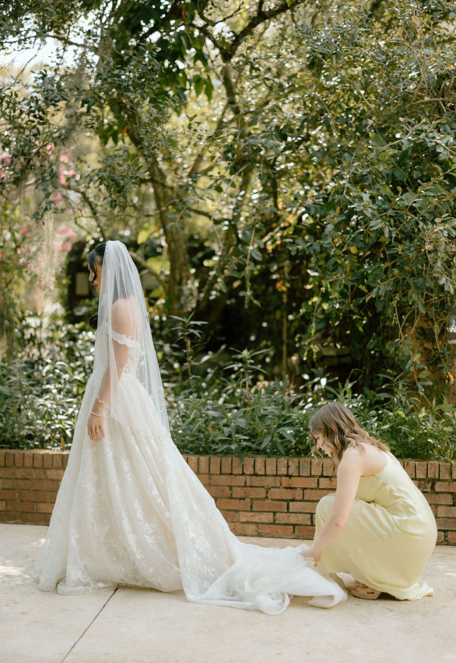 A bride in a white lace gown with long veil has her dress adjusted outdoors in a garden setting.
