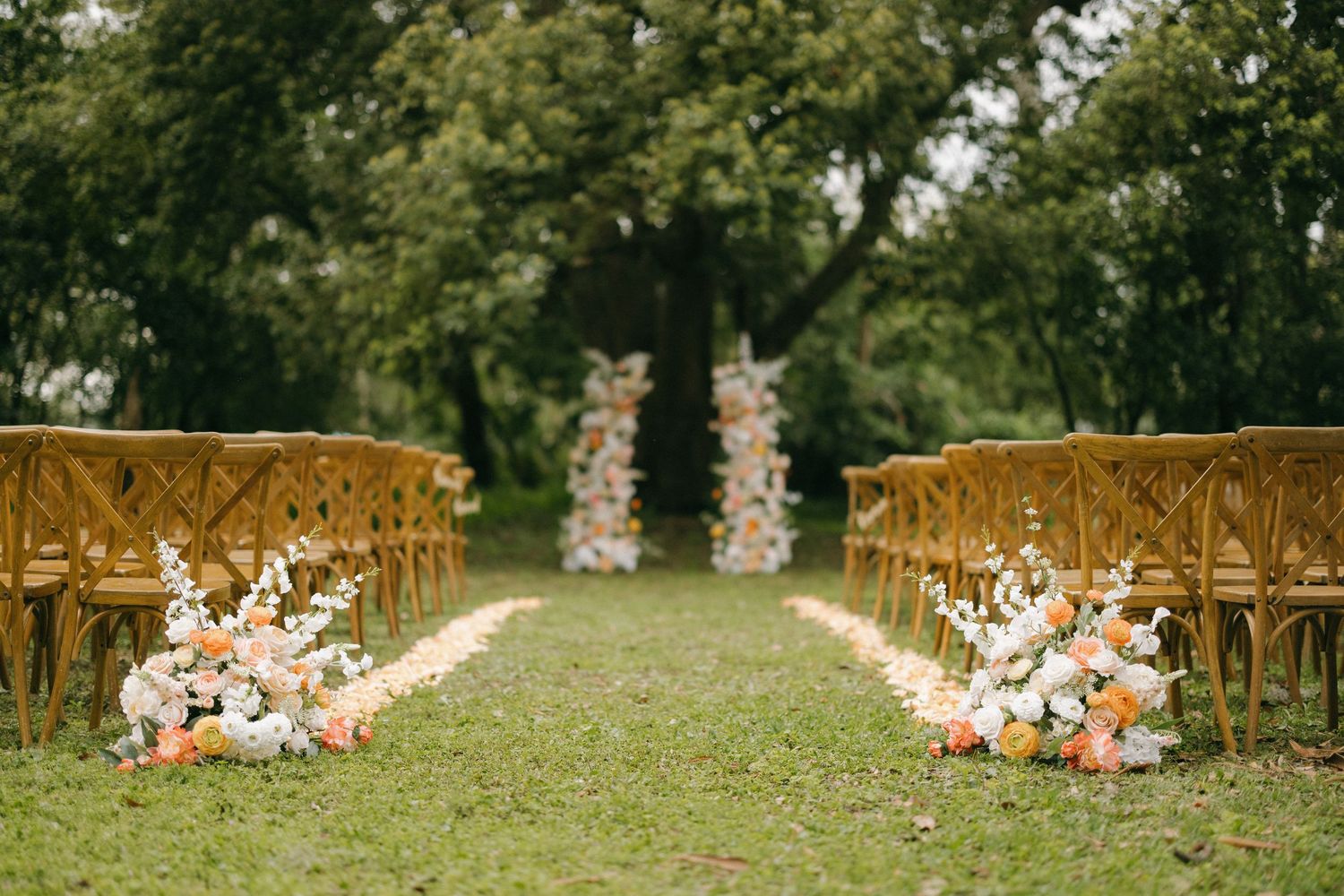 A whimsical styled wedding with wooden ceremony chairs arranged in rows on a grassy lawn with floral arrangements lining the aisle.