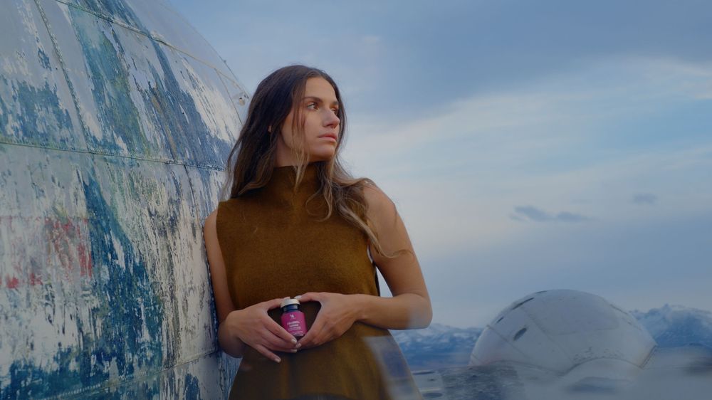 A person in brown clothing stands on a scenic mountain summit overlooking a vast mountain range and cloudy sky.