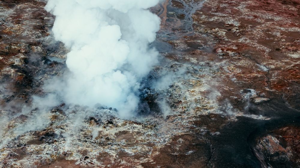 Aerial view of a geothermal landscape with steaming hot springs and reddish-brown mineral deposits.