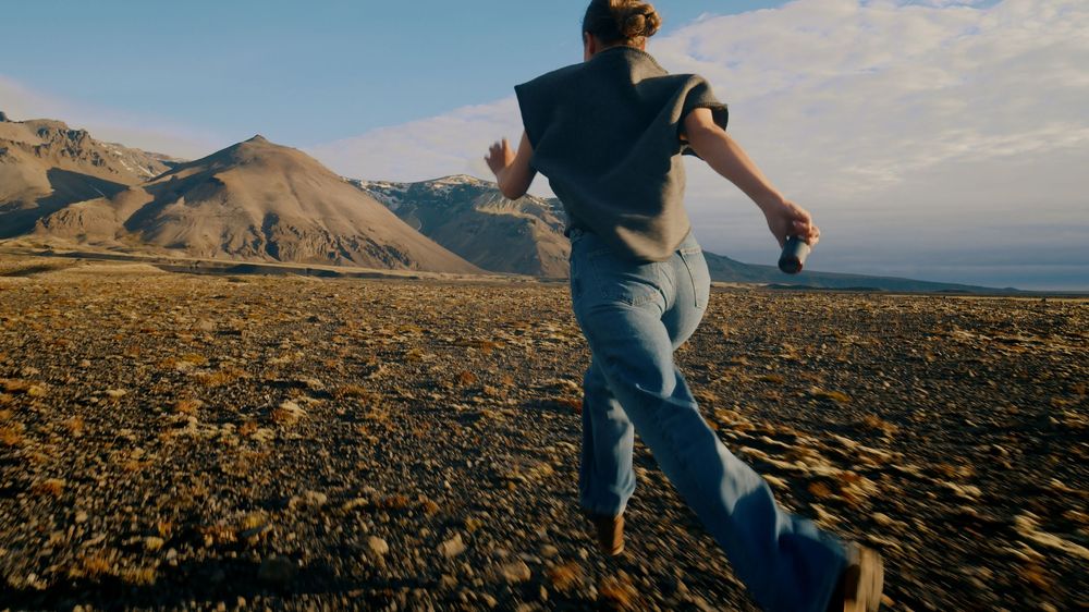 Person walking across rocky mountain terrain at sunset with dramatic panoramic views of rugged landscape.