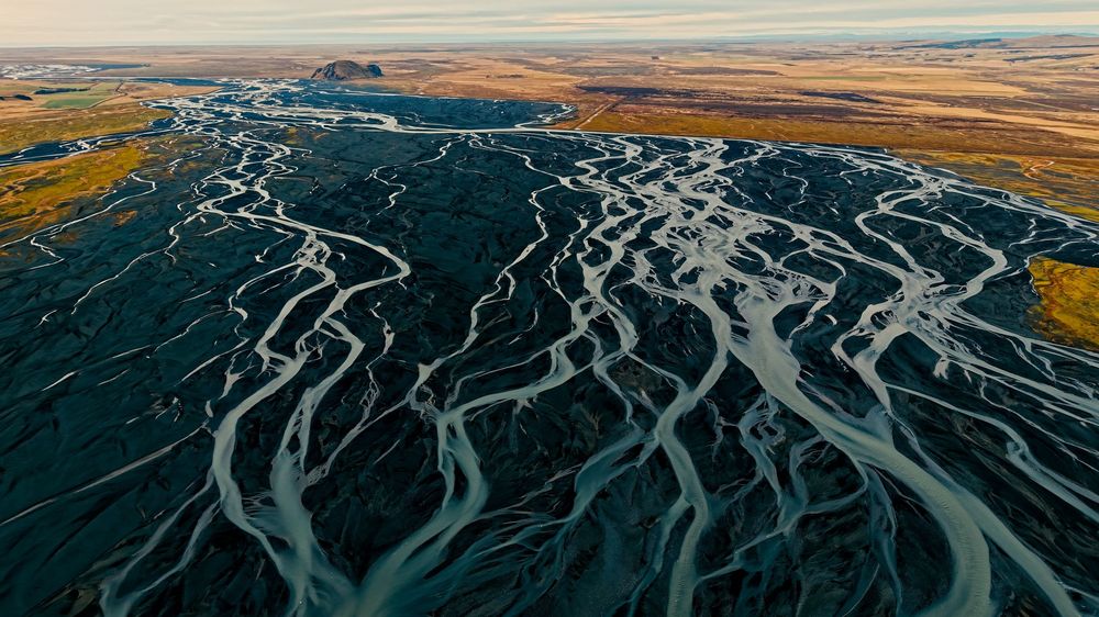 Aerial view of glacial river channels forming intricate braided patterns across dark volcanic landscape in Iceland.