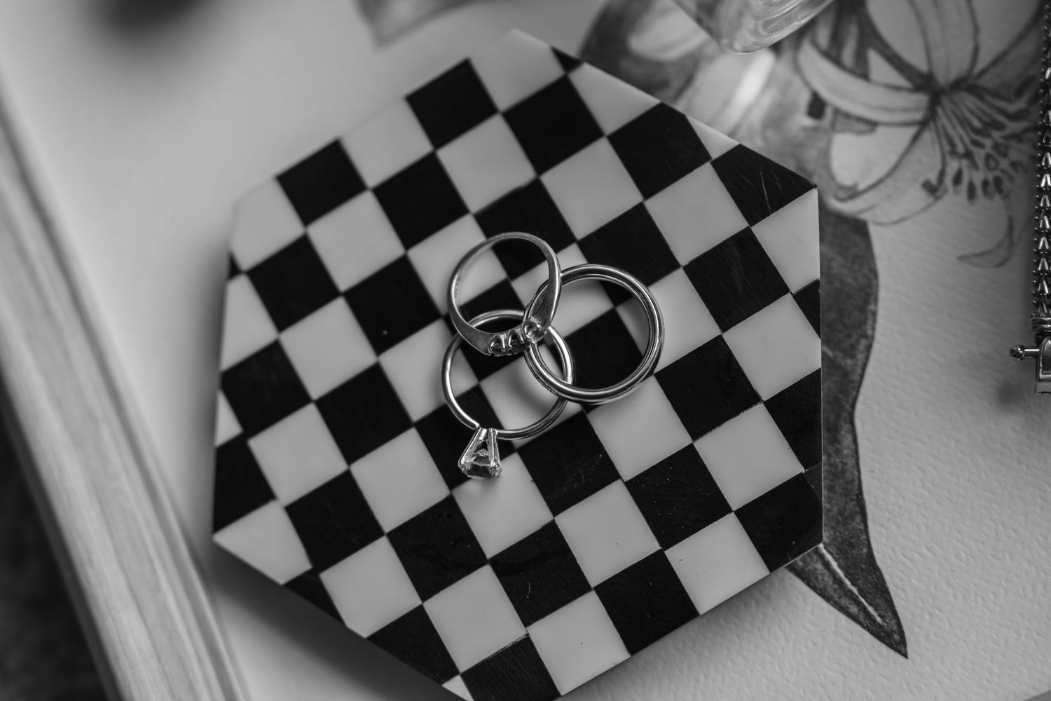 Two wedding rings rest on a black and white checkered pattern surface in an artistic monochrome photograph.