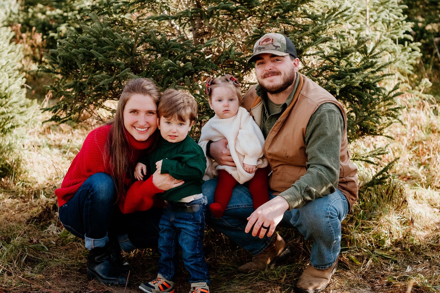 Family of four poses together in front of evergreen trees.