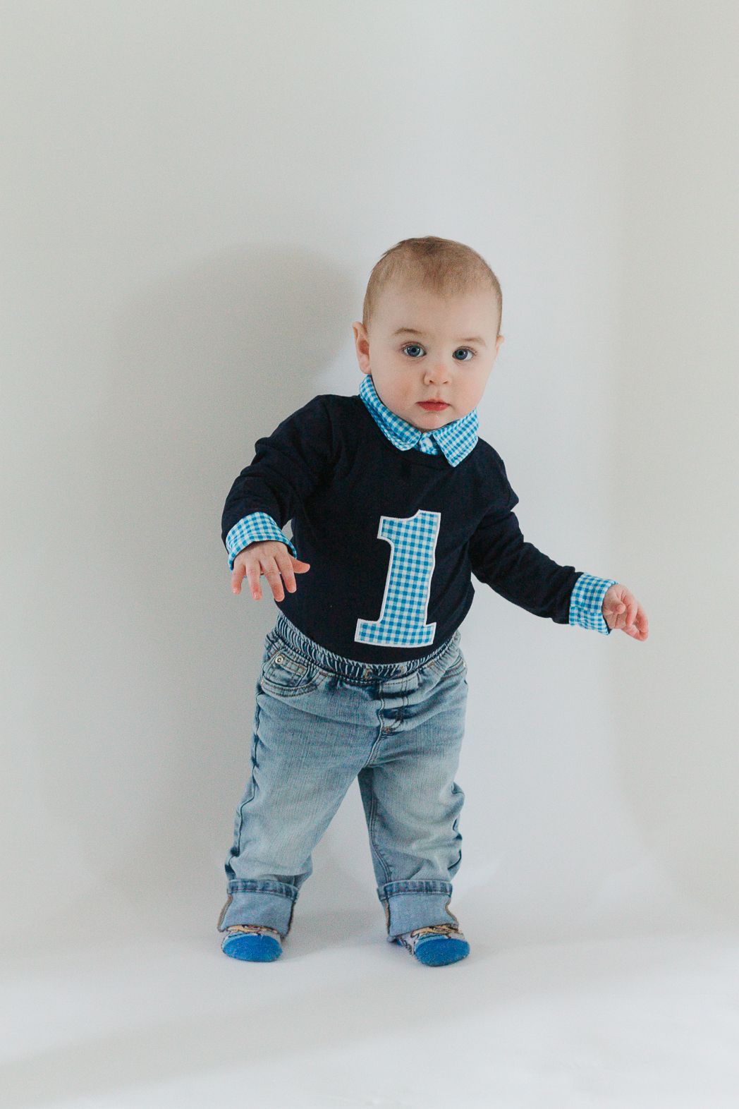 Baby wearing a navy sweater with blue number one and jeans stands against white background.