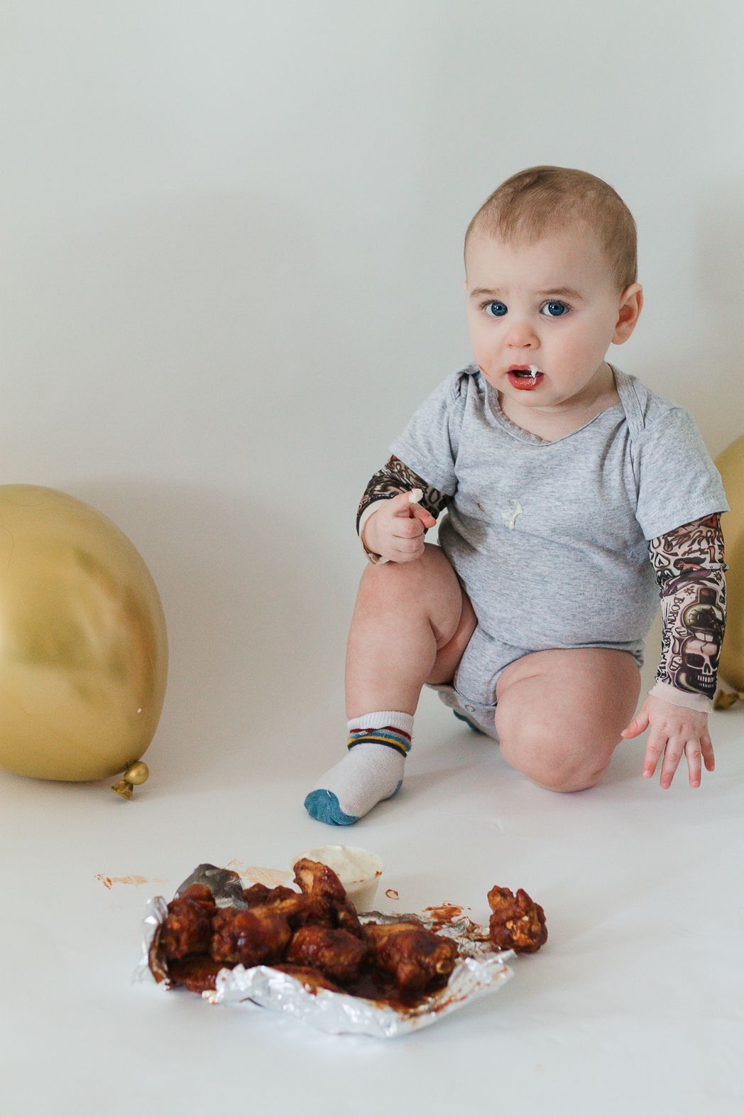 Baby in grey onesie sits next to chicken wings and gold balloon on white background.