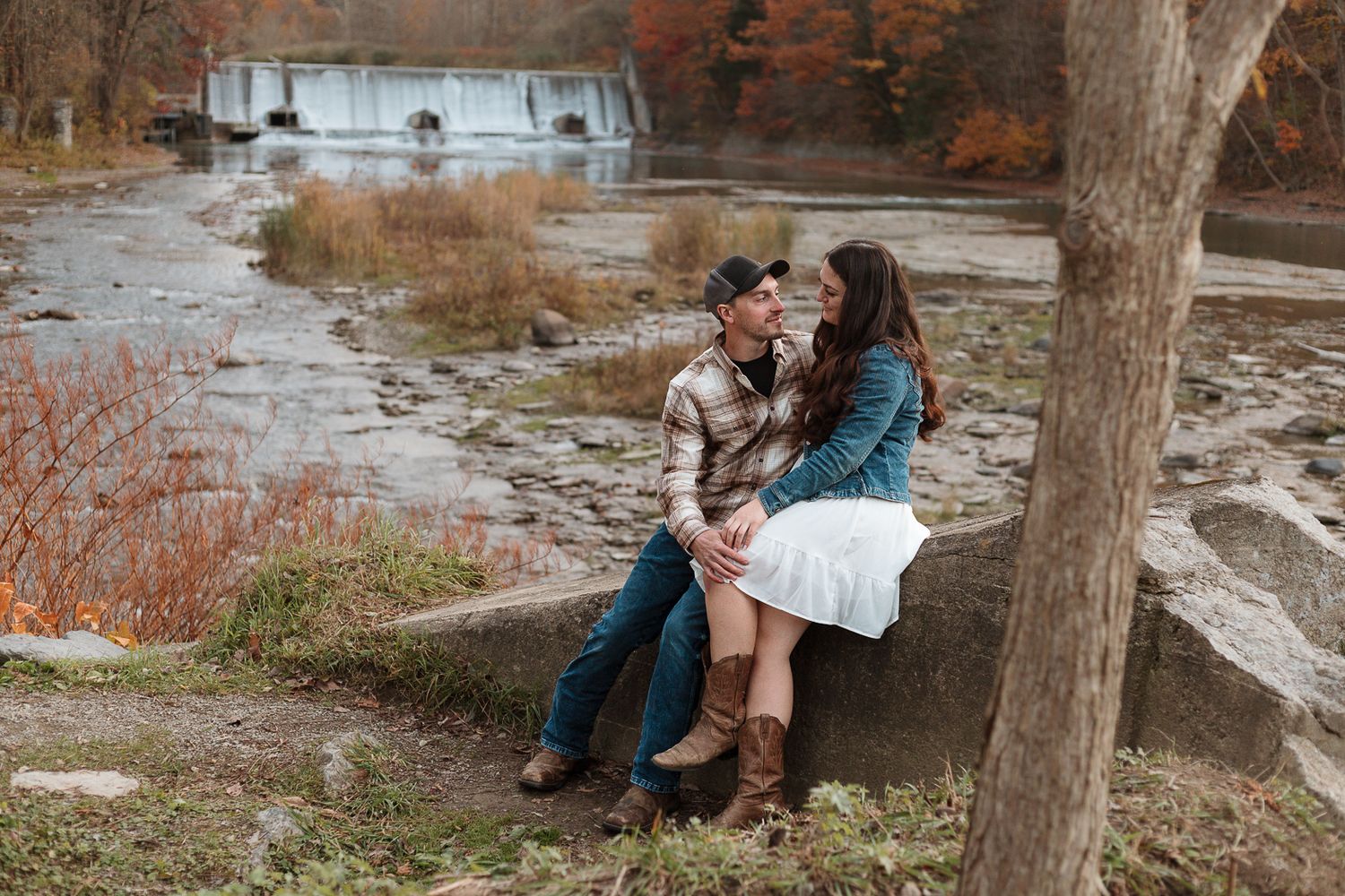 A couple shares tender moments against a backdrop of autumn trees and water.