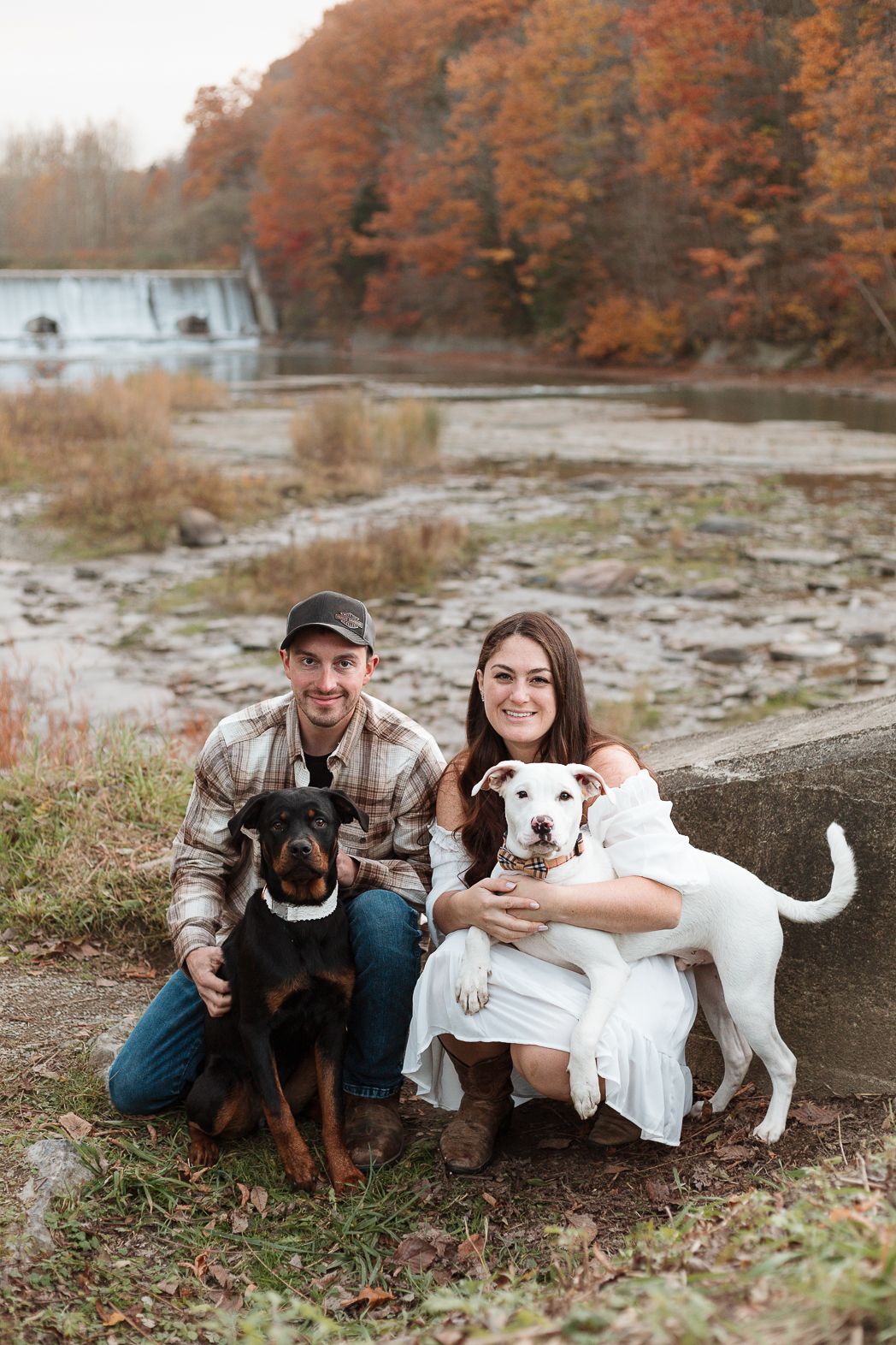 A couple sits with their two dogs on rocky terrain surrounded by fall foliage.
