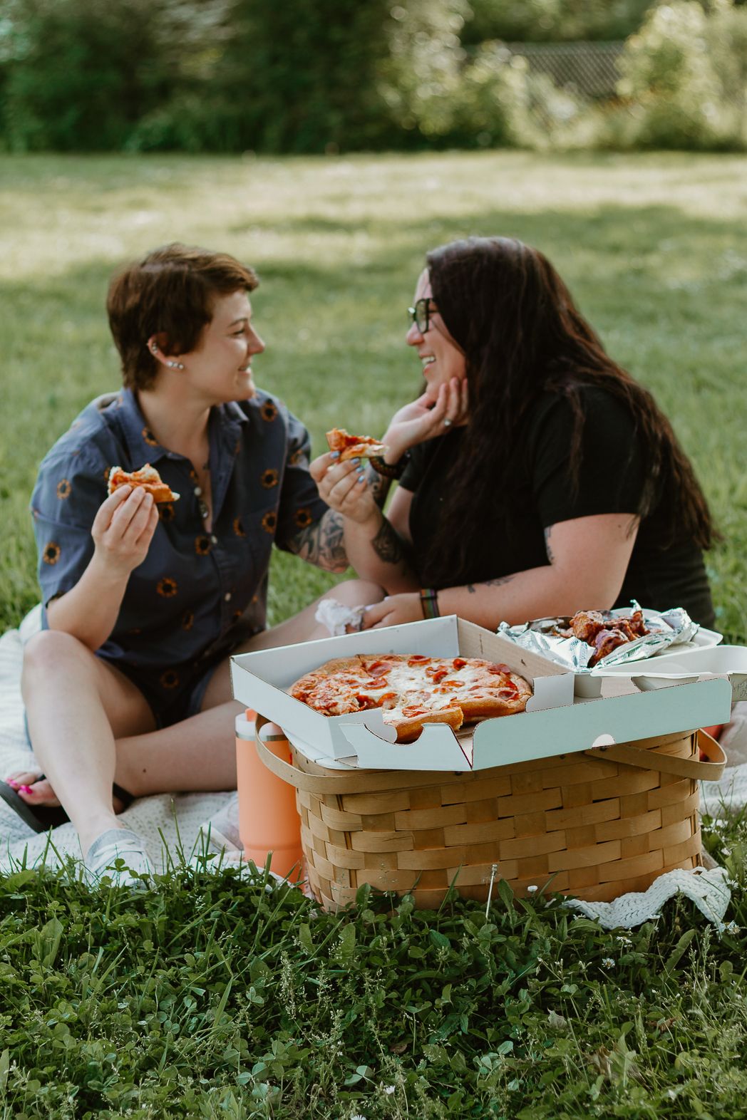 Friends having a picnic on grass with woven basket full of snacks.
