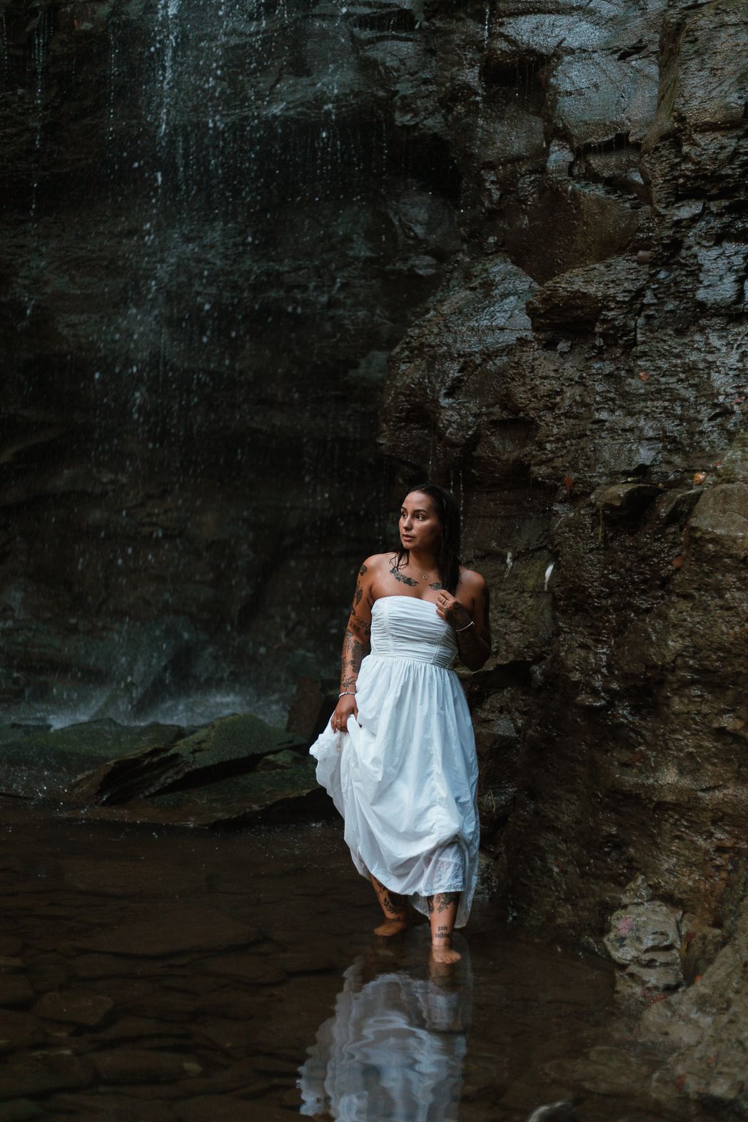 Person in white dress wades through dark water near rocky cliff face.