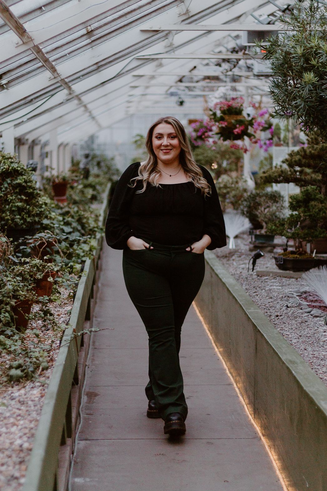 Woman in black outfit walking through greenhouse with plants and flowers.