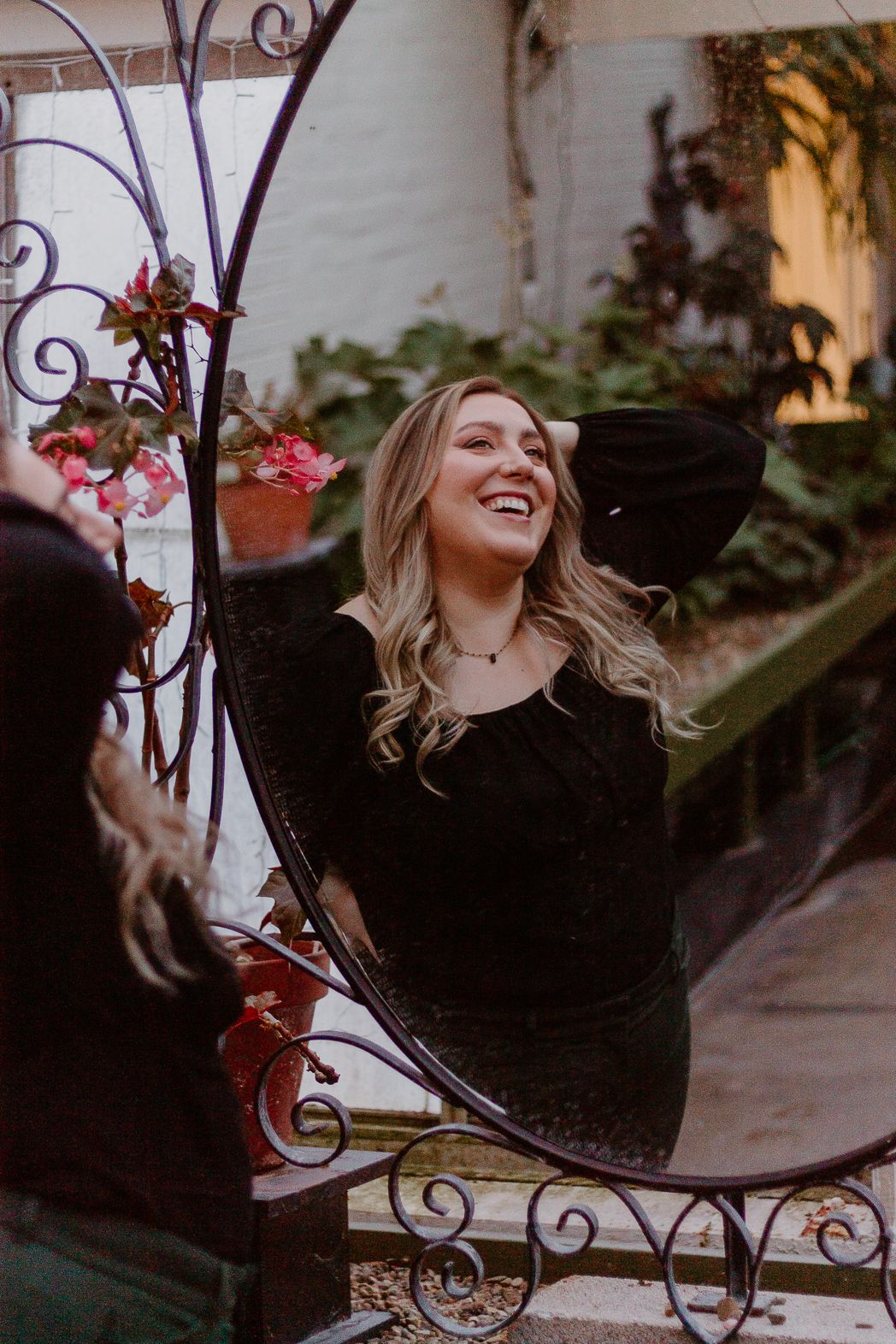 Woman in black dress laughing by decorative iron gate with pink flowers.