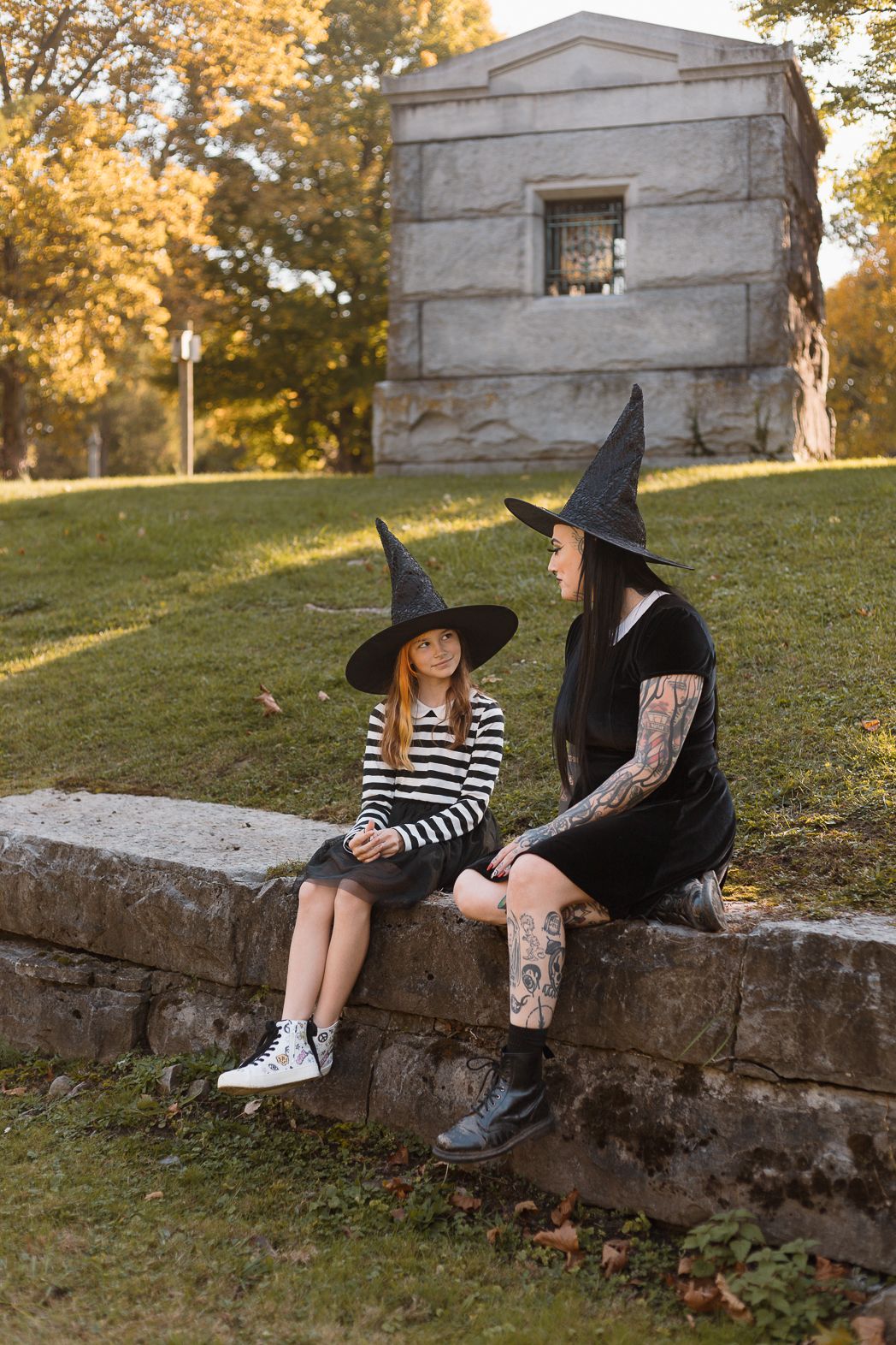 Two people in witch costumes sit on a stone wall with a historic building in the background.