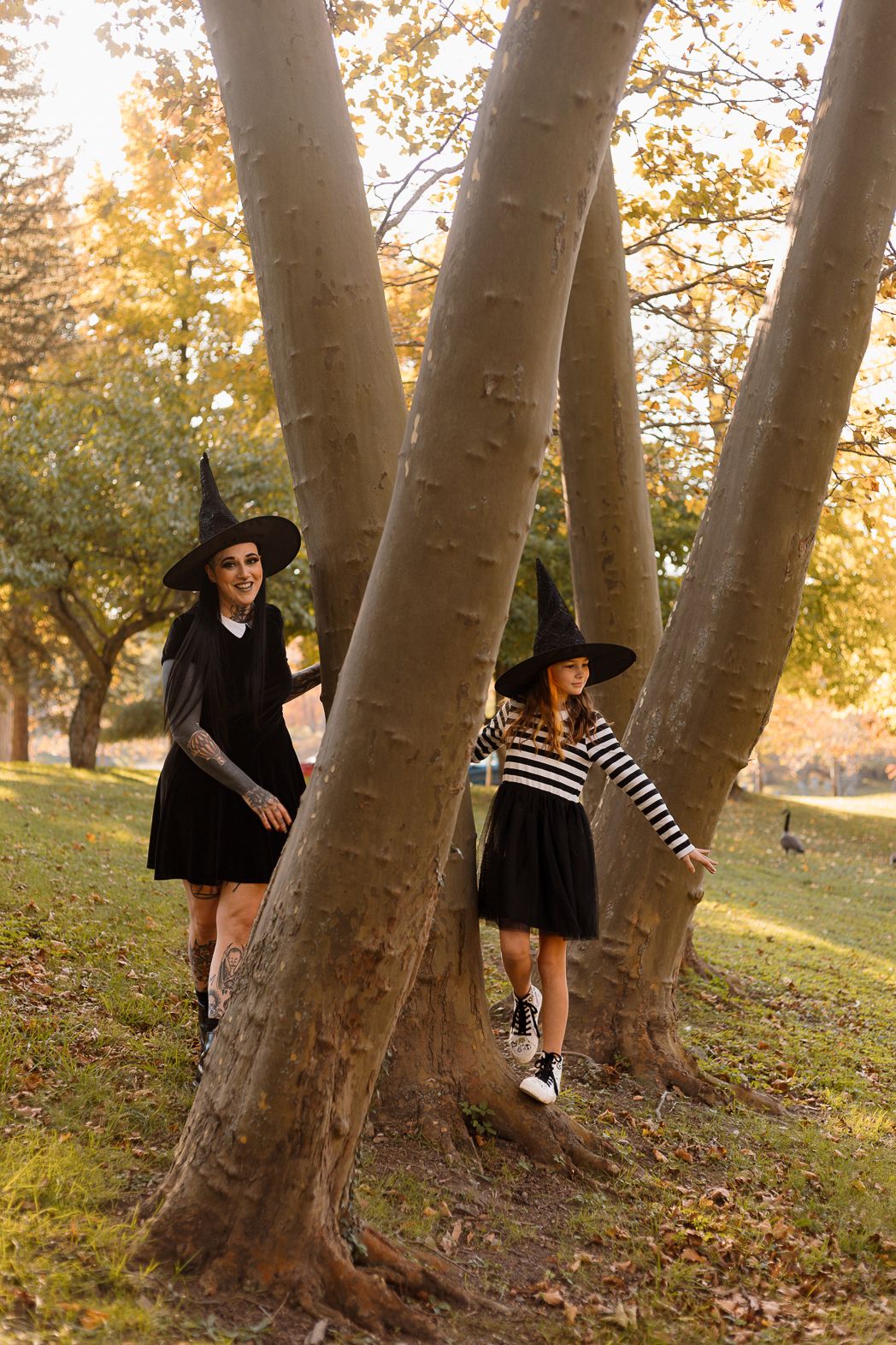 Two people in witch costumes play among large tree trunks in an autumn park.