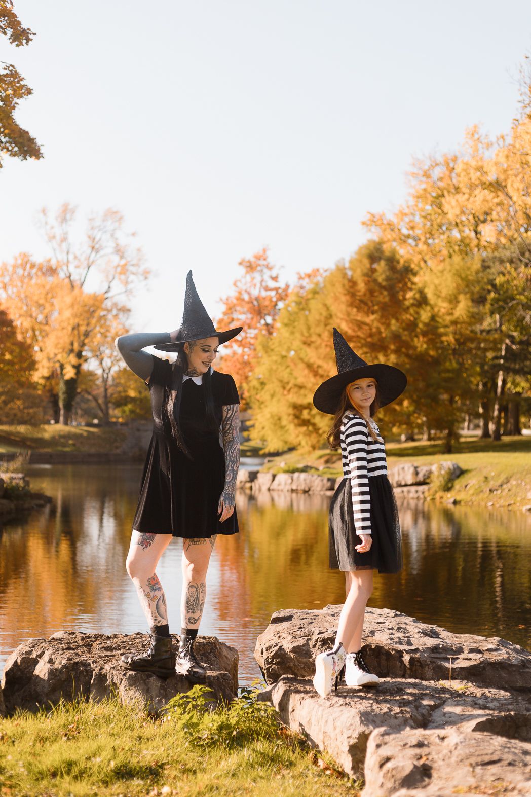 Two people in witch costumes pose on rocks by a pond during autumn.