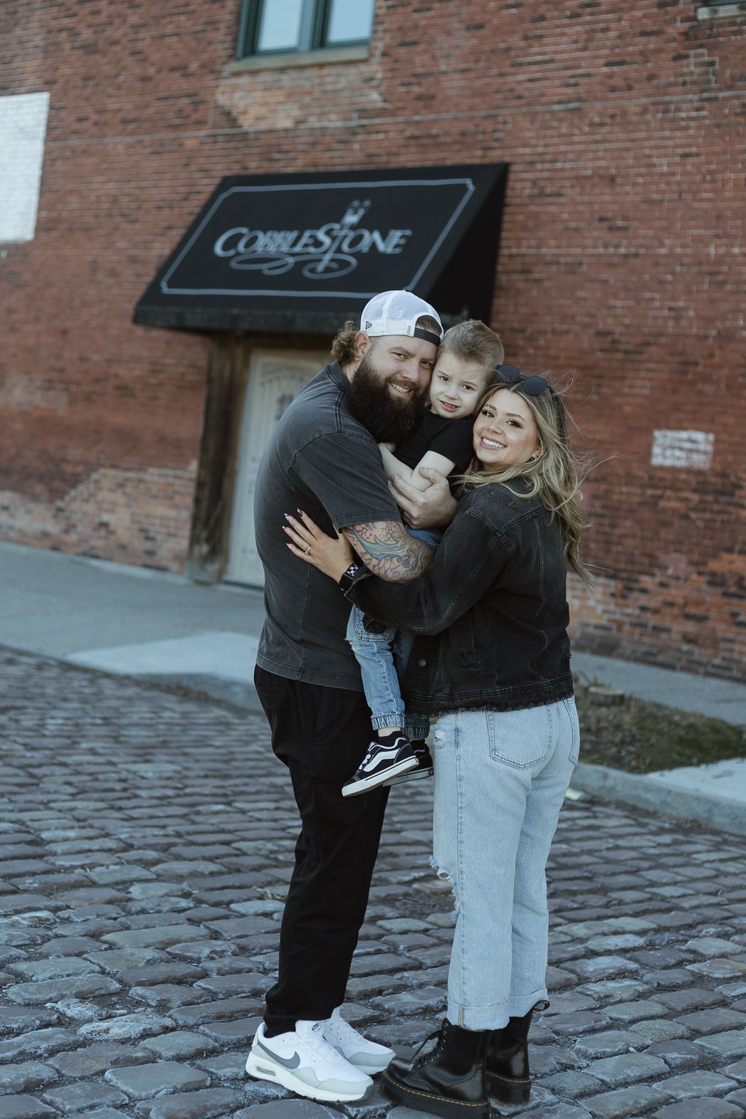 Two young children in casual winter outfits hug and play together on a cobblestone street near a brick building.