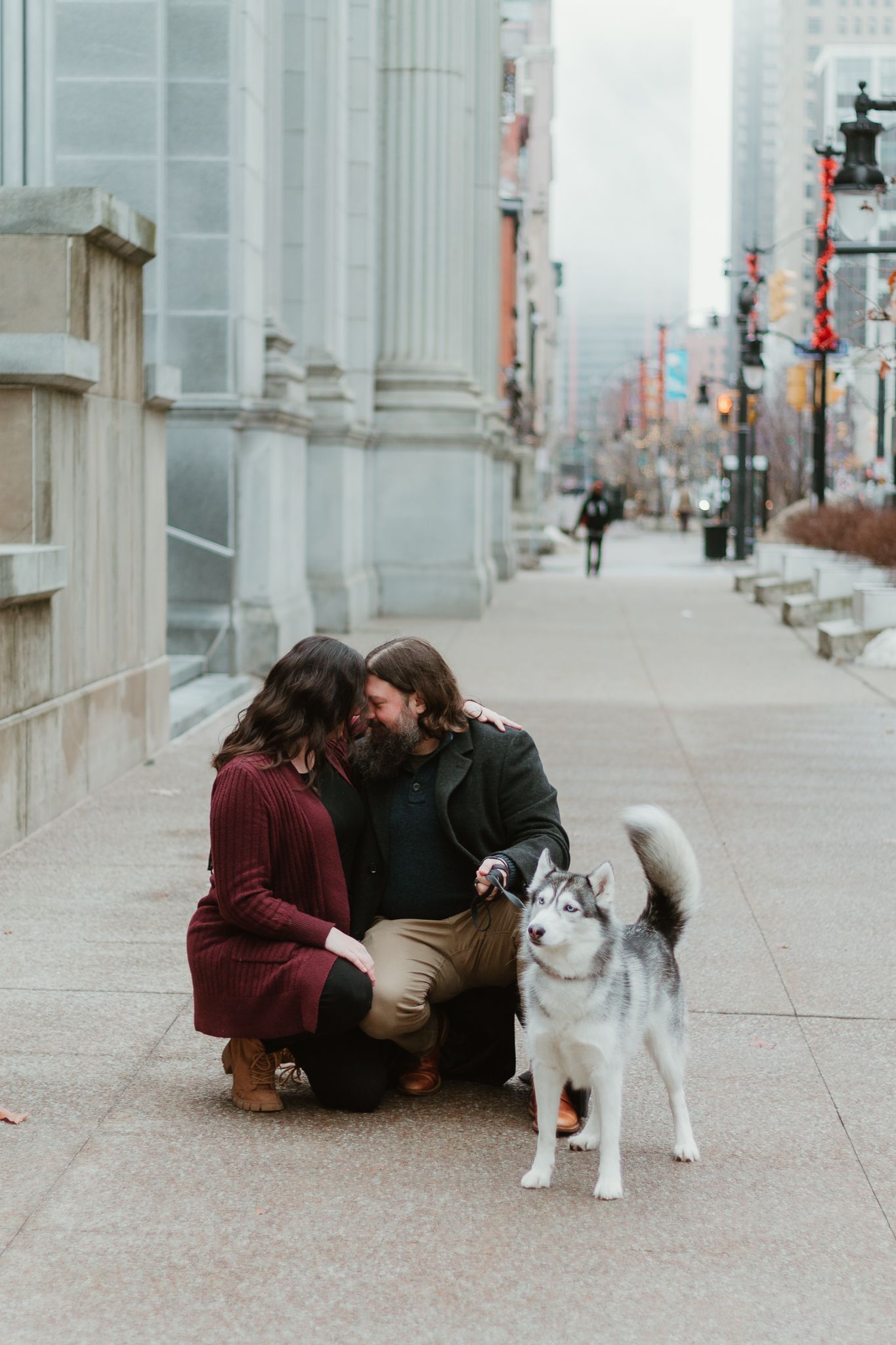 Couple crouching with their Husky dog on city sidewalk.