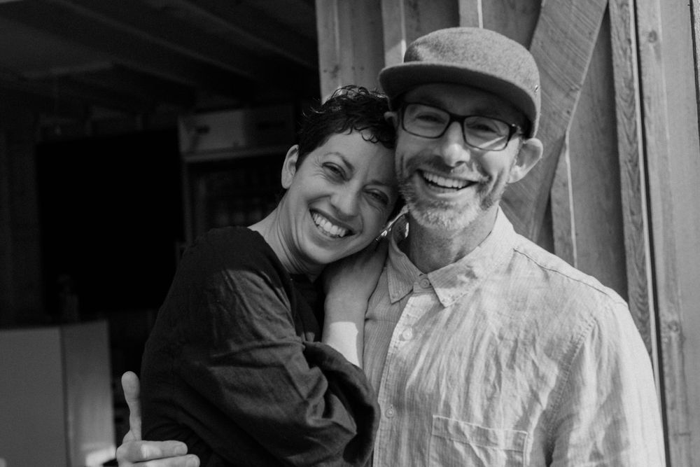 A black and white portrait of two people smiling and embracing by a wooden wall.