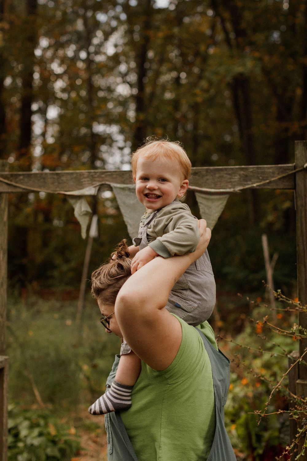 A joyful toddler sits on shoulders against a wooded autumn background.