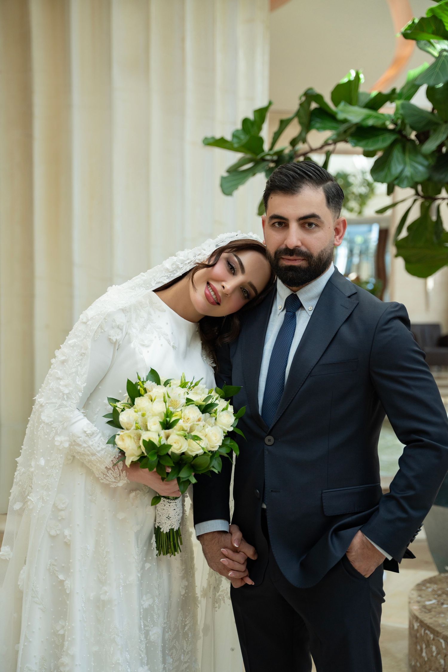 A newlywed couple posing at their wedding, with the bride in white lace holding a white bouquet and groom in dark suit.