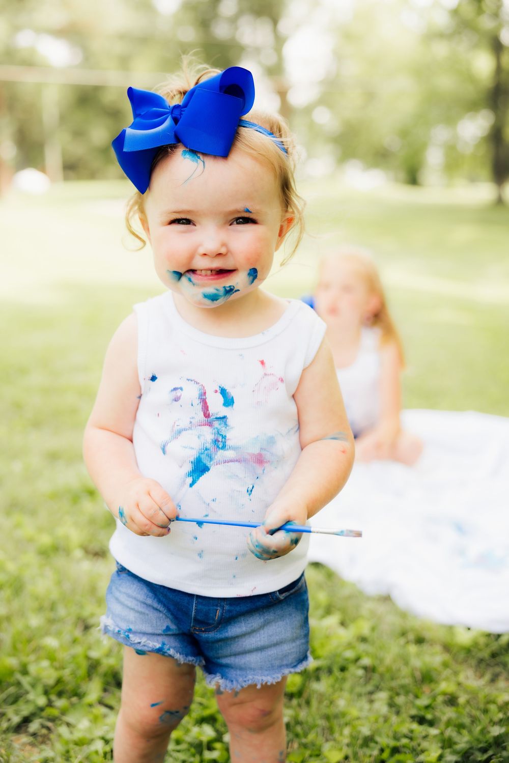 A toddler in denim shorts and white top plays with blue paint outdoors.