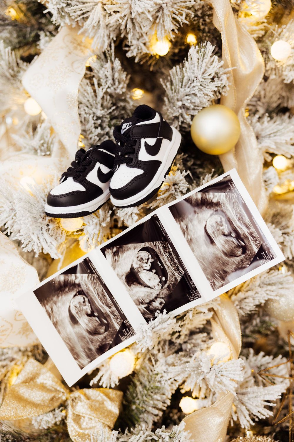 Ultrasound photos and baby shoes displayed on white flocked Christmas tree branches with gold ornaments.