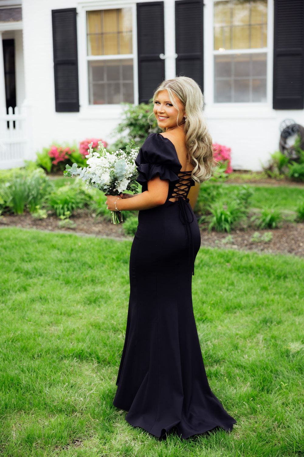 A prom mini session featuring a senior in an elegant black dress poses with white flowers on a lawn.