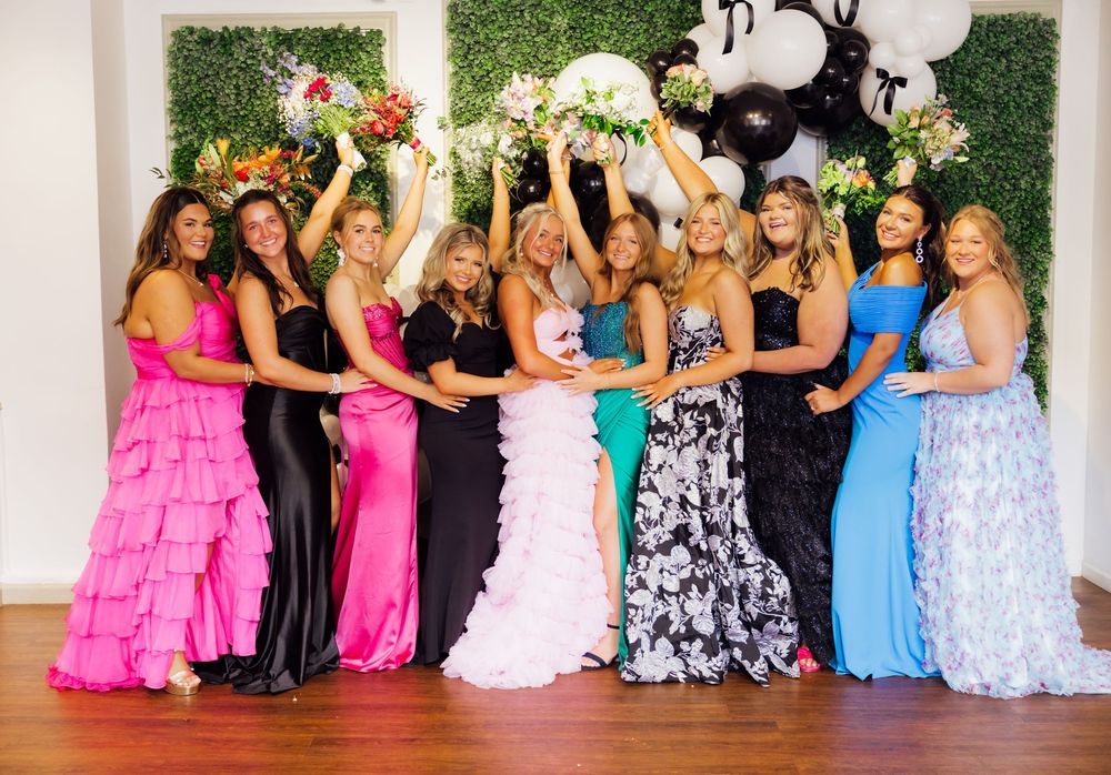 A group prom mini session photo of people wearing colorful formal dresses posed against a green hedge wall backdrop.