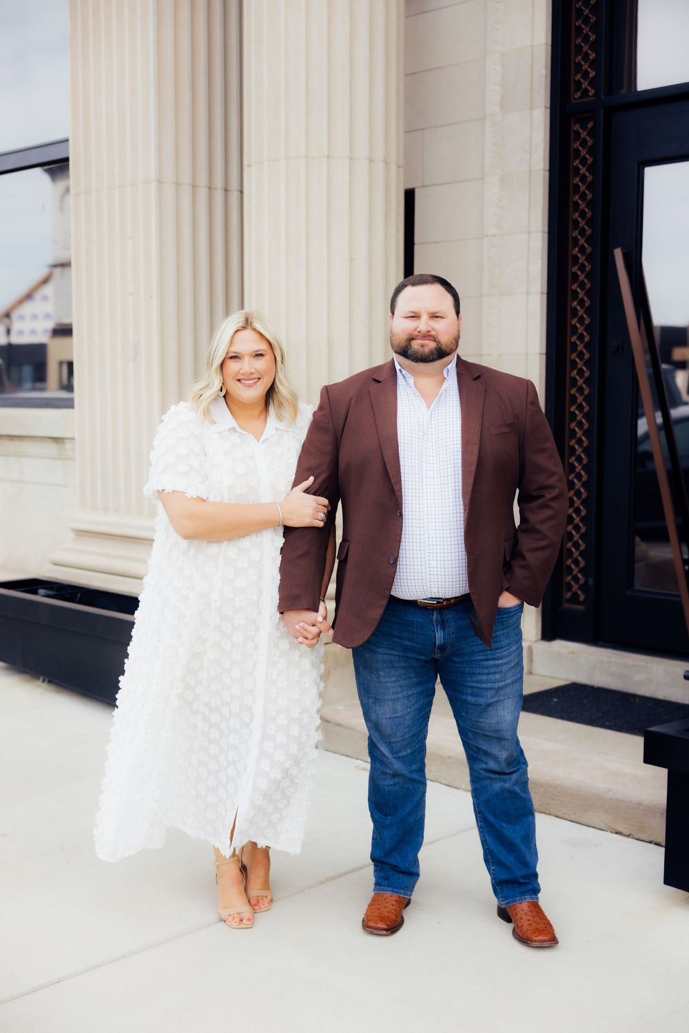Couple session posing in front of white columns, wearing brown blazer and white dress.